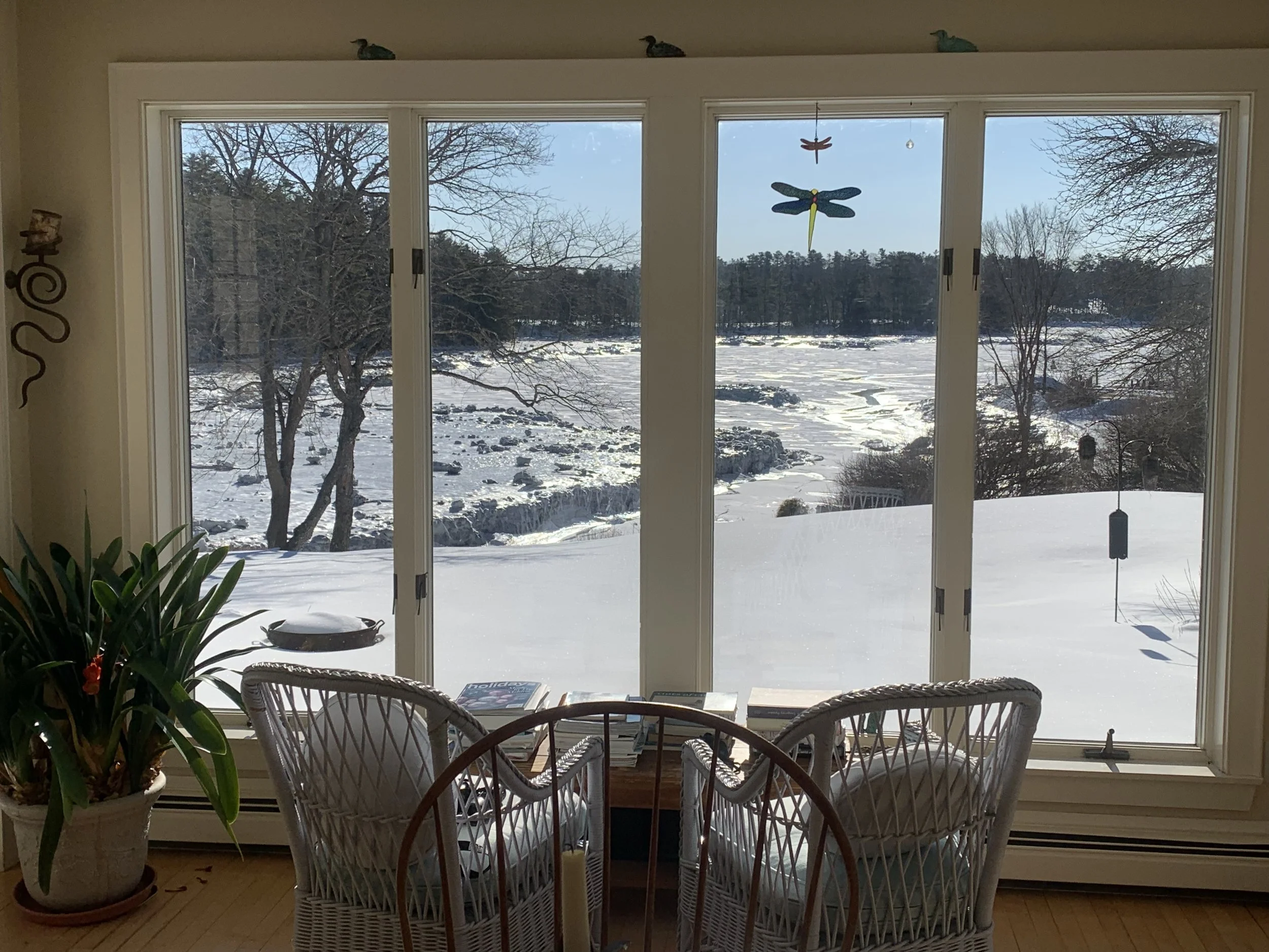 A room with a large window showing a snowy outdoor landscape, with leafless trees, a frozen river or pond, a small table with chairs, a potted plant, and hanging decorations inside.