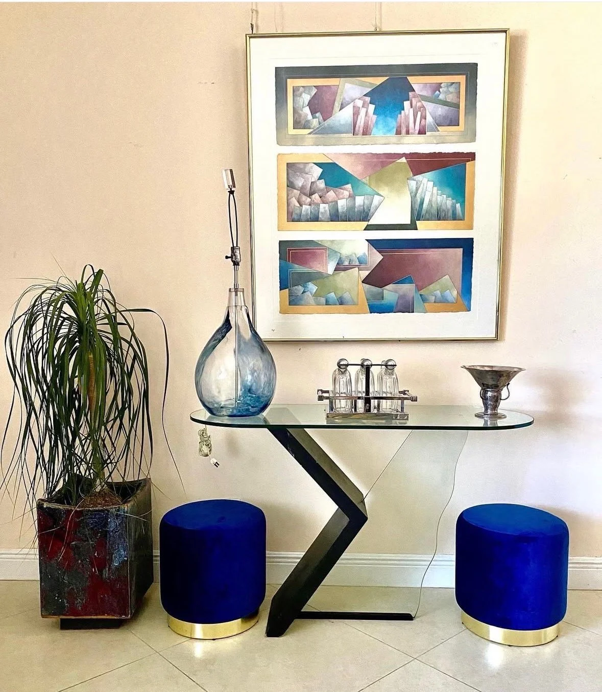 Decorative glass table with a zigzag black base, two blue velvet round stools with gold bases, a tall plant in a red marble planter, and a framed abstract geometric art piece on a beige wall.