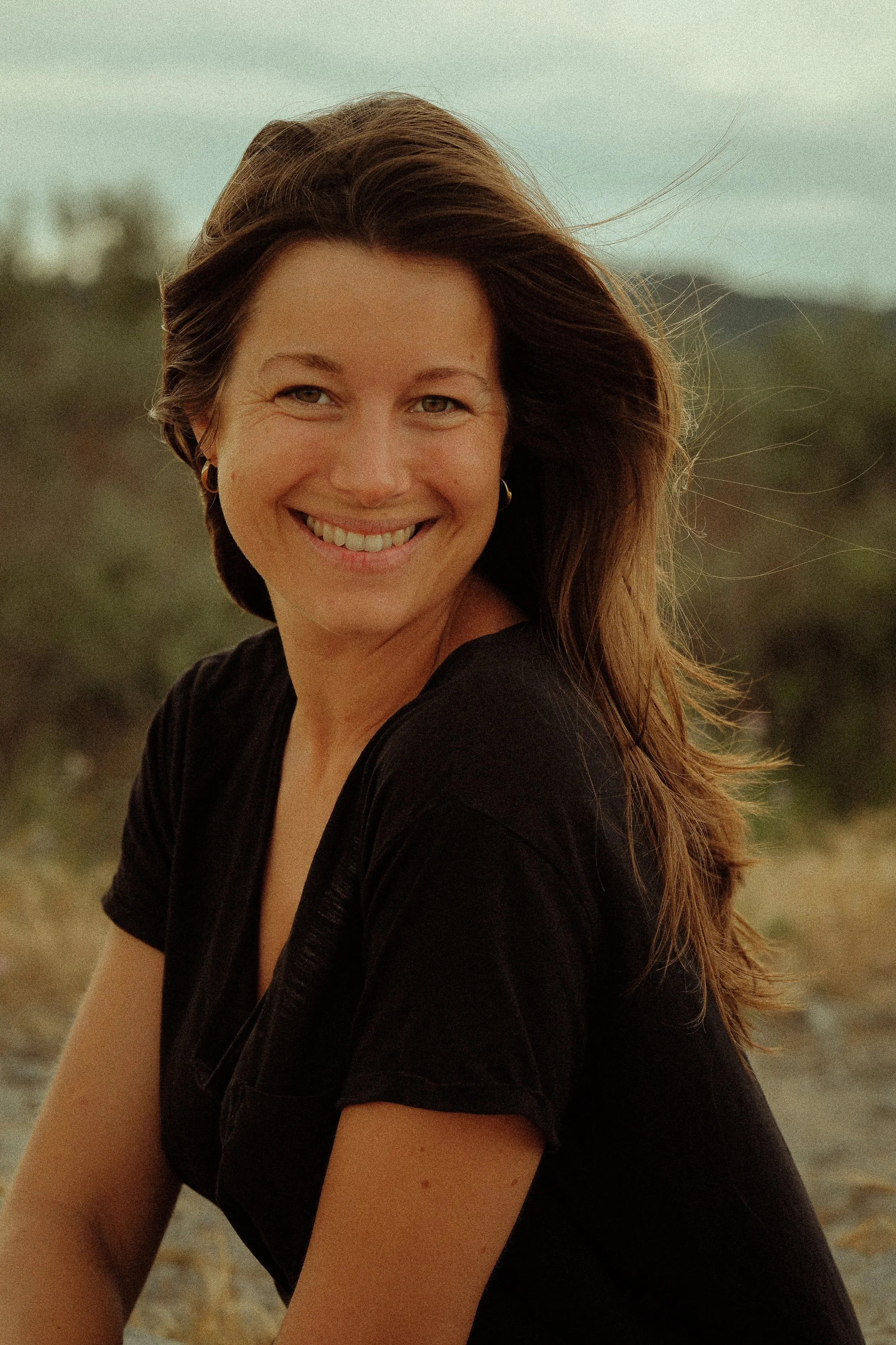 Portrait de Tiphaine Deuff. Une femme souriante avec des cheveux longs et bruns, portant un haut noir, assise à l'extérieur avec un fond flou de nature.