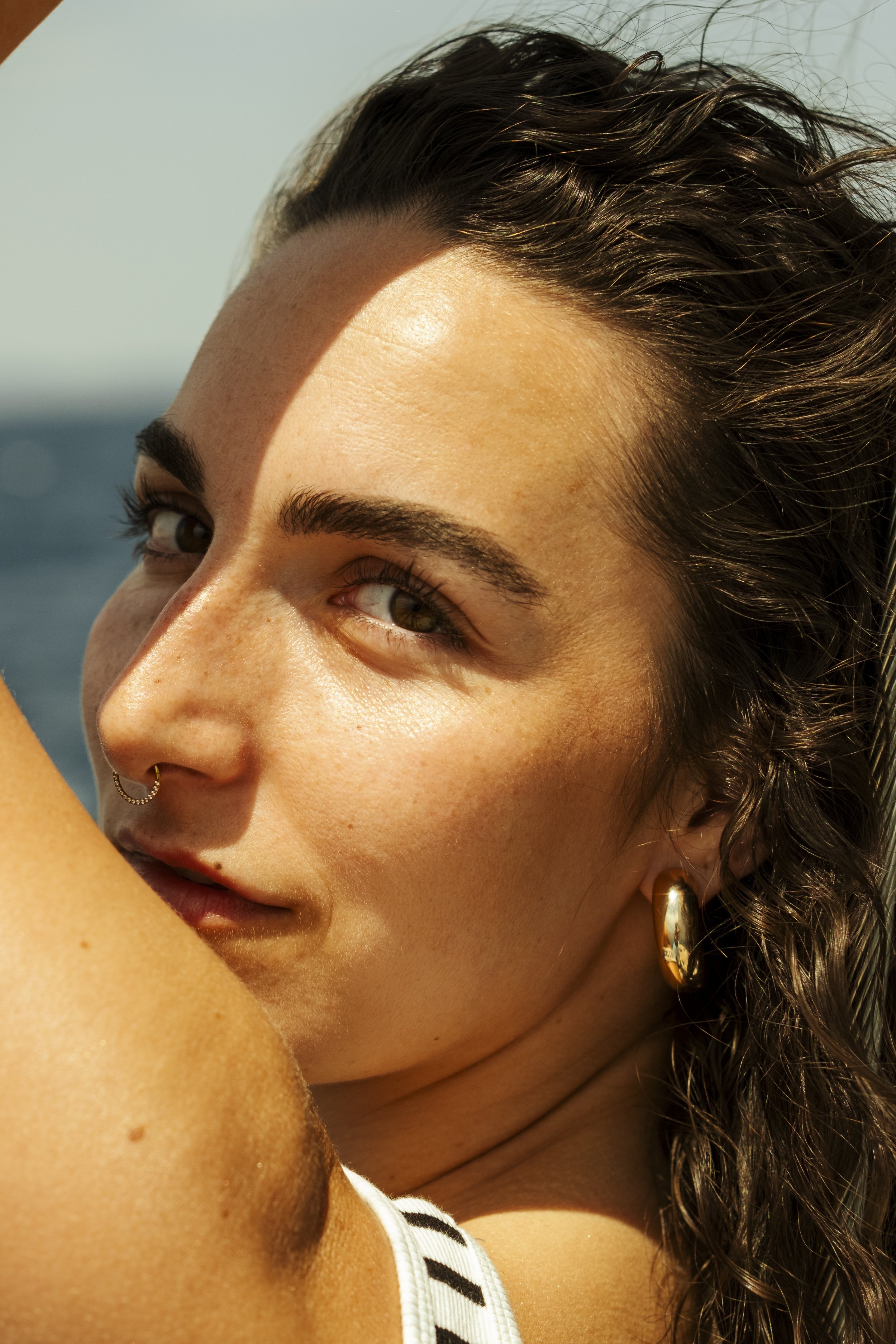 Une jeune femme avec des cheveux bruns, maquillée, portant un bijou en or, regarde en direction de la droite. La photo est prise en gros plan, avec une lumière naturelle.