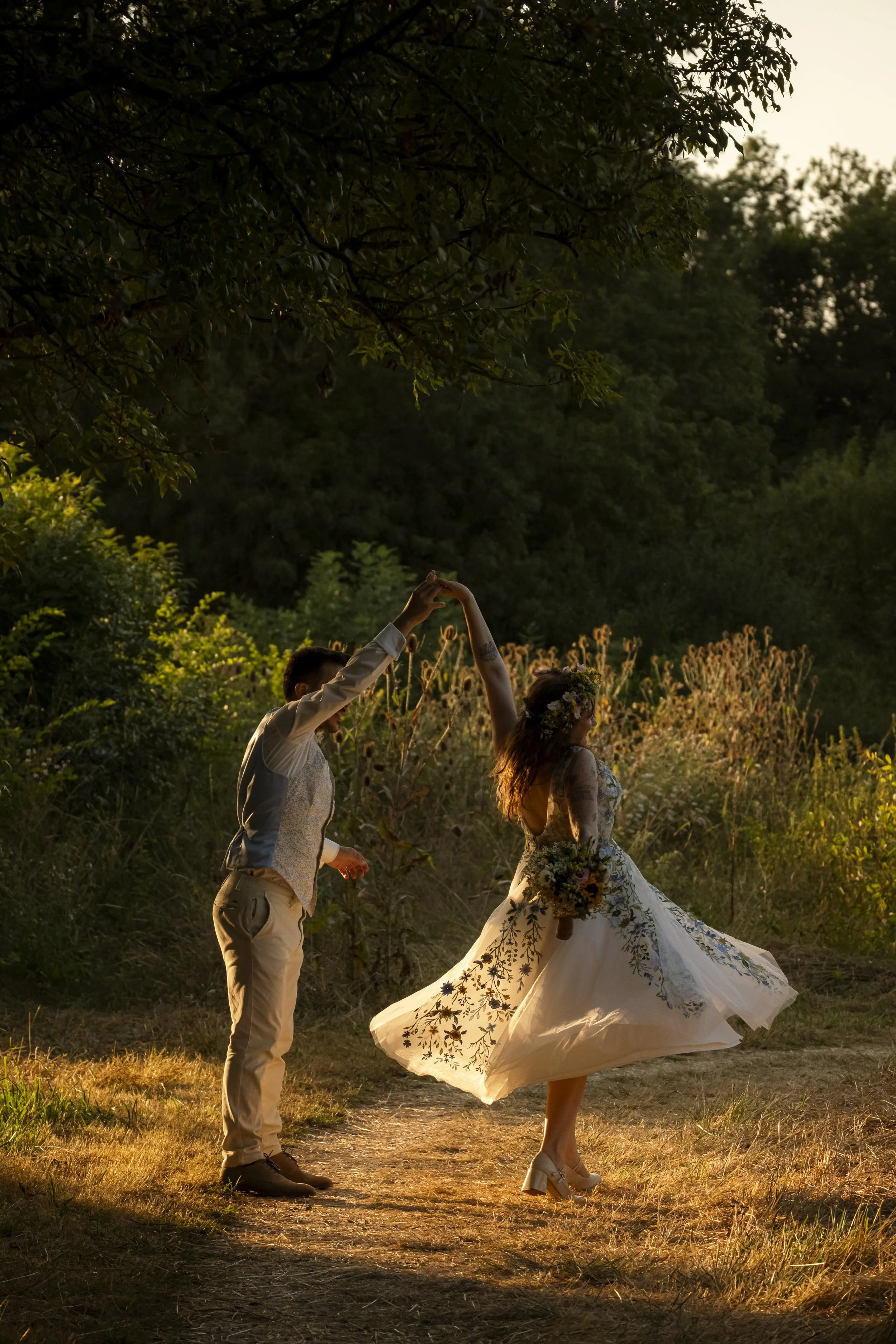Un couple danse lors d'une cérémonie en plein air au coucher du soleil, l'homme porte un costume clair, la femme porte une robe de mariage fluide avec des motifs floraux.