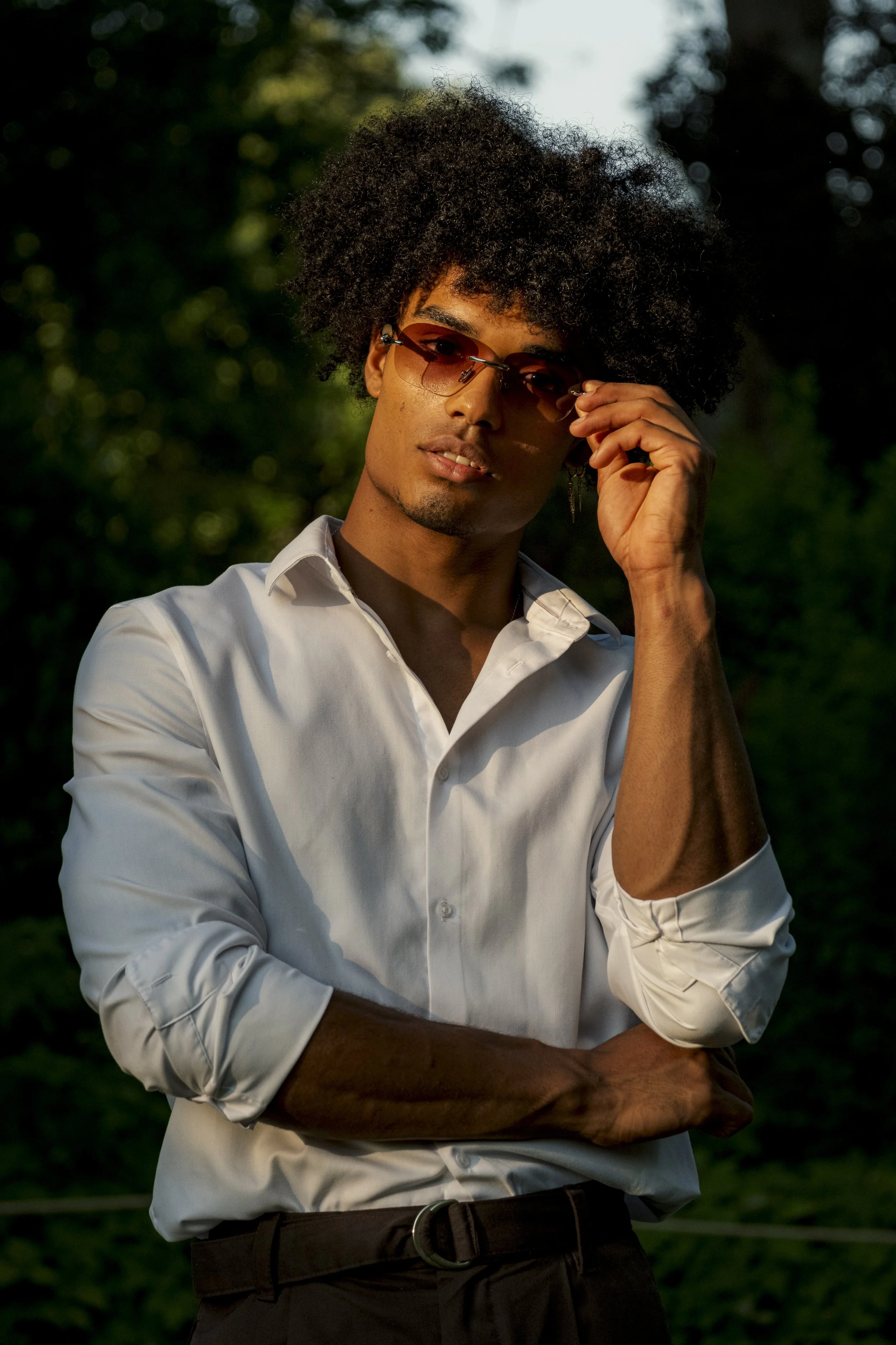 Jeune homme portant des lunettes de soleil, chemise blanche, dans un environnement extérieur verdoyant, avec un éclairage tamisé, souriant légèrement, croisé les bras.