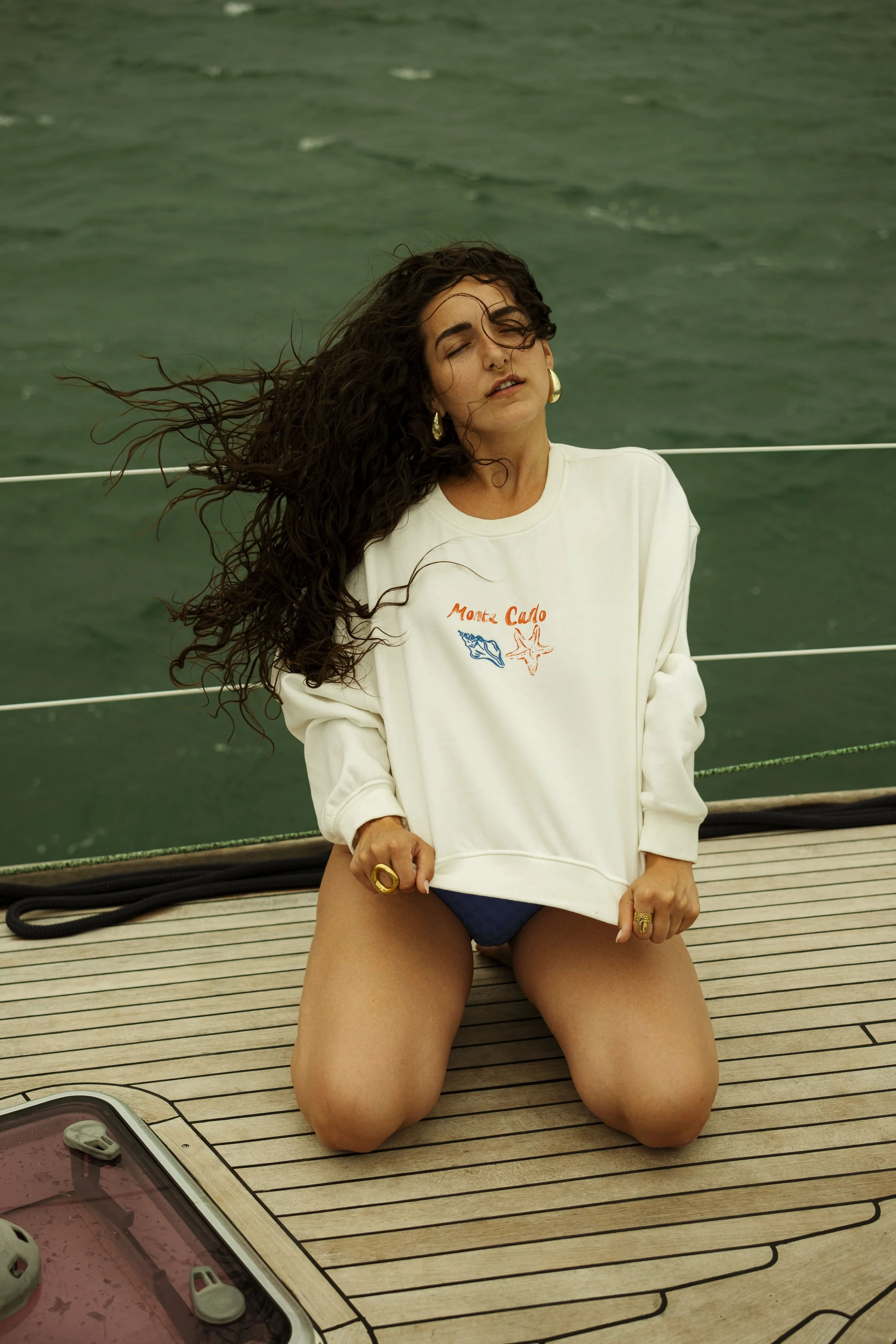 Une femme avec des cheveux bouclés longs, portant un sweat blanc avec 'Monte Carlo' et des illustrations, posant sur un bateau avec une mer en arrière-plan.