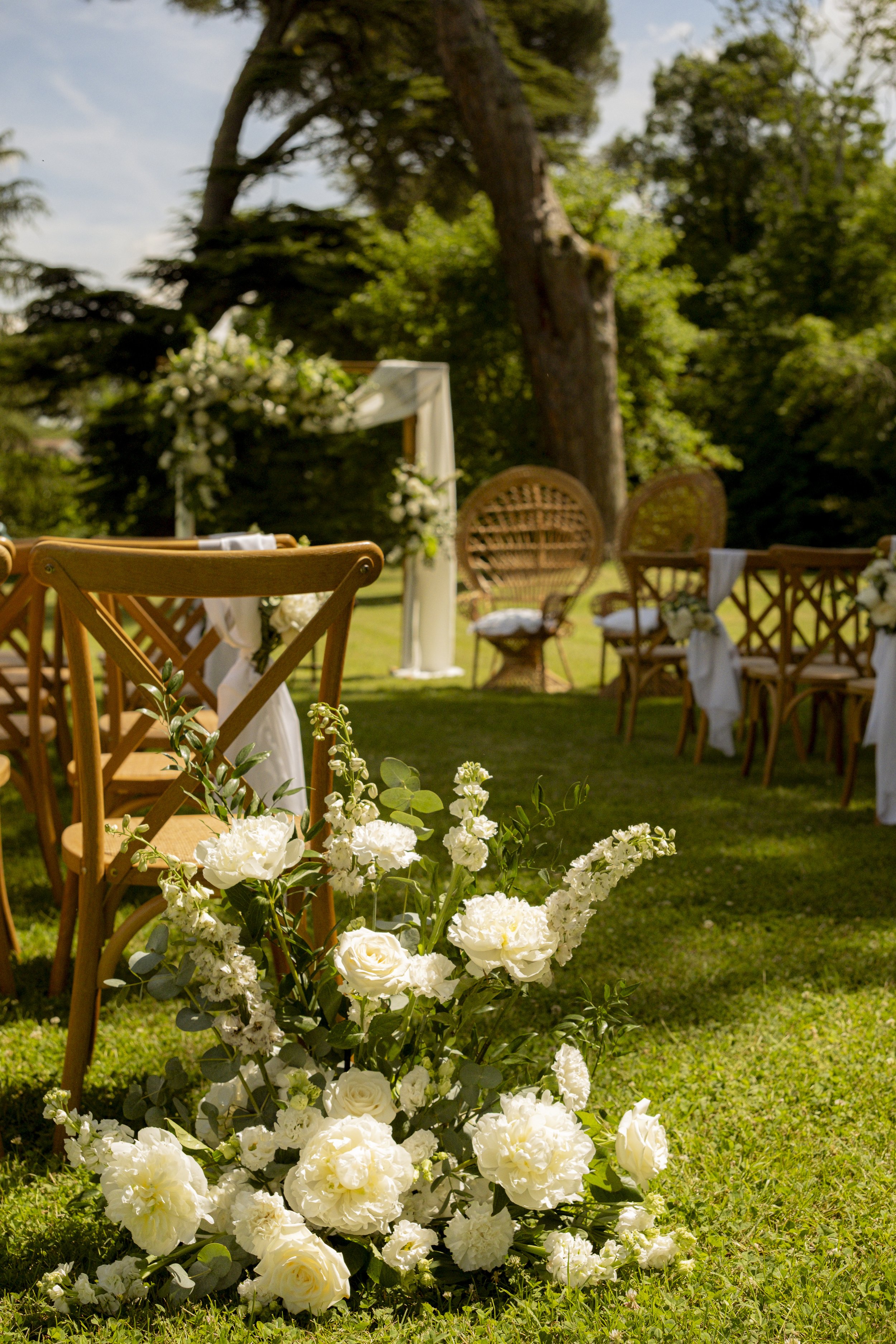 Décor de mariage en plein air avec fleurs blanches, chaises en bois, et un arbre en arrière-plan sous un ciel ensoleillé.