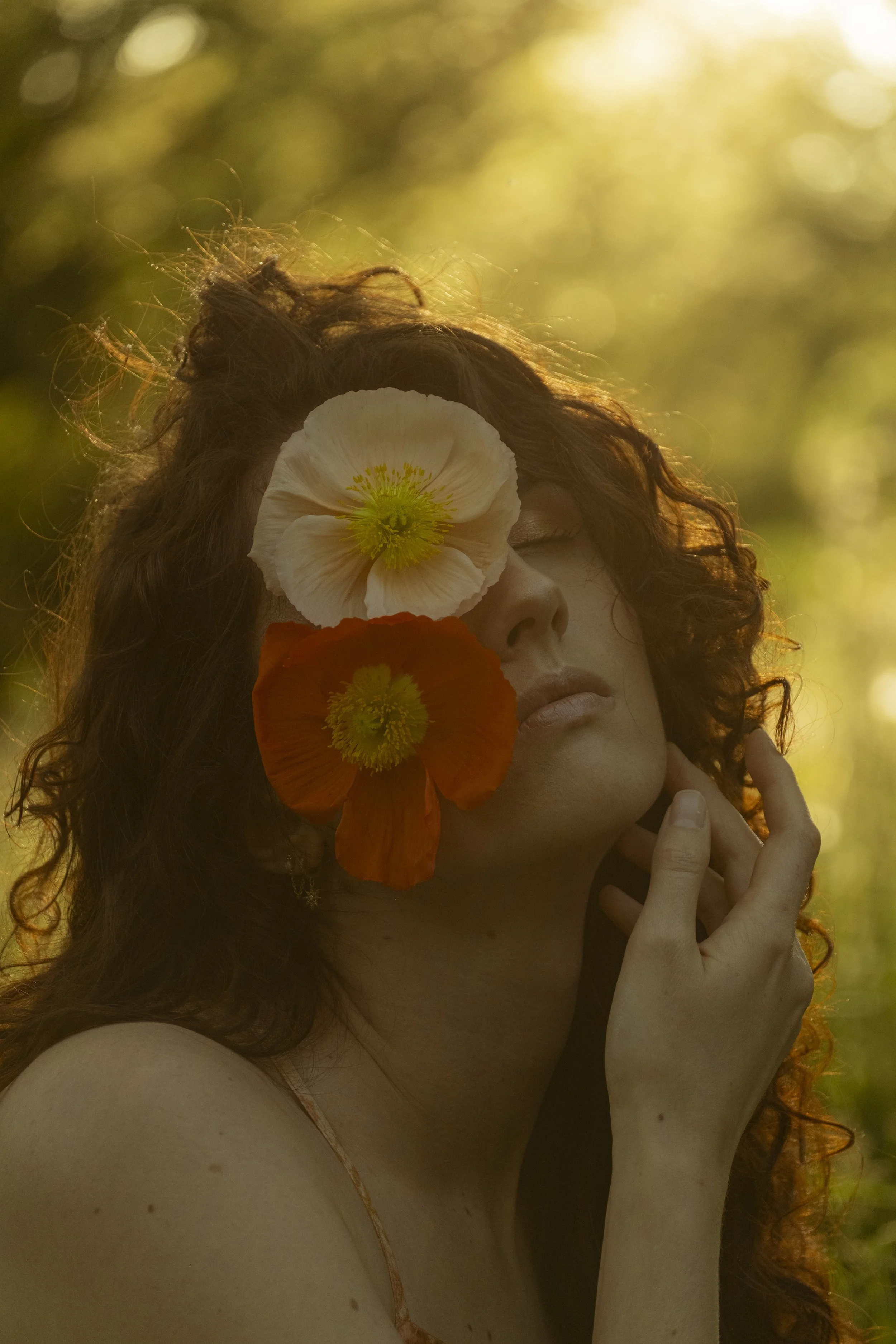 Portrait d'une femme aux cheveux bouclés, avec des fleurs orange et blanches près du visage, éclairée par une lumière chaude en arrière-plan.