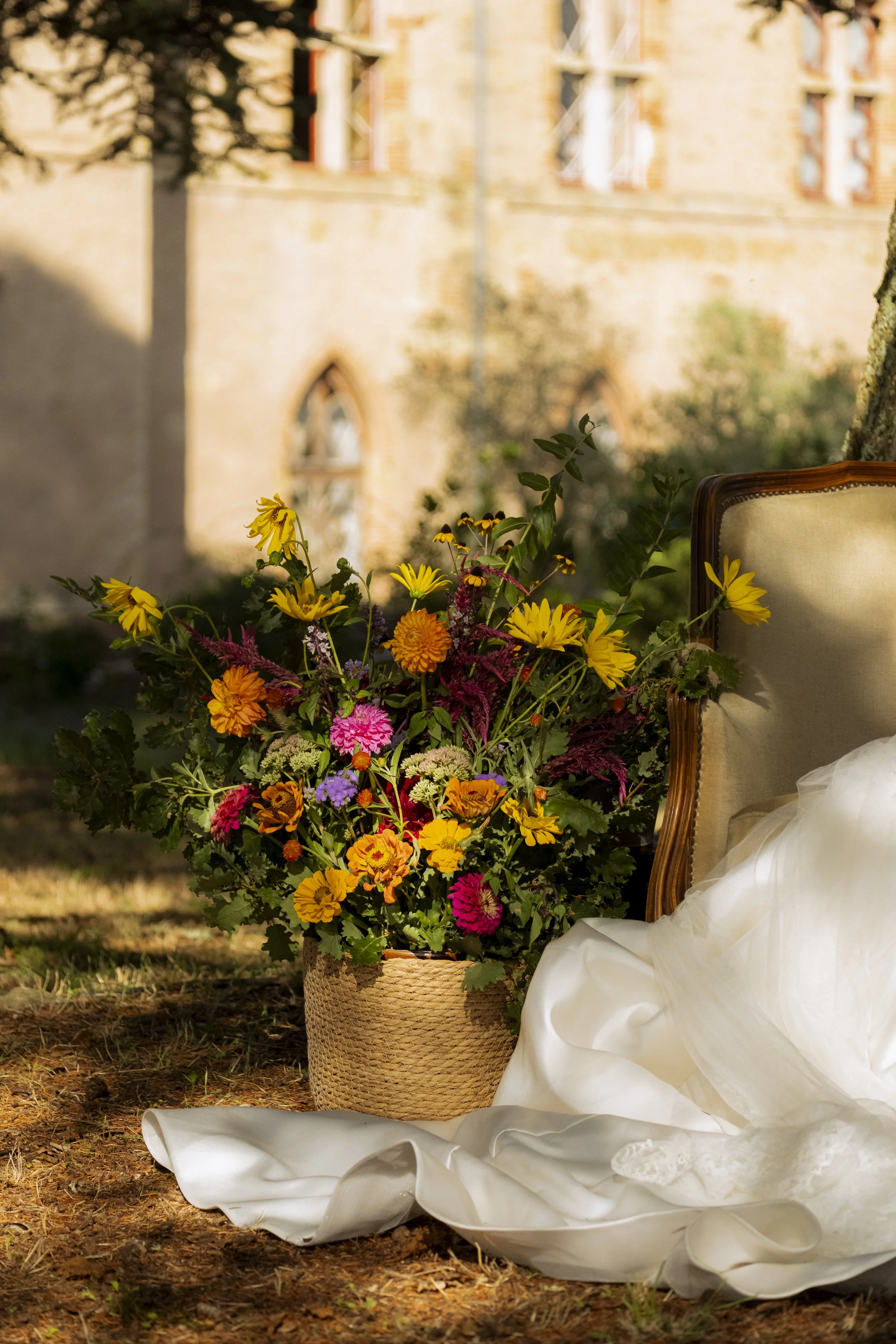 Un panier de fleurs colorées, un fauteuil ancien en bois avec un tissu beige, et des vêtements blancs en tissu léger posés à côté, dans un cadre extérieur avec des bâtiments en pierre et des arbres.