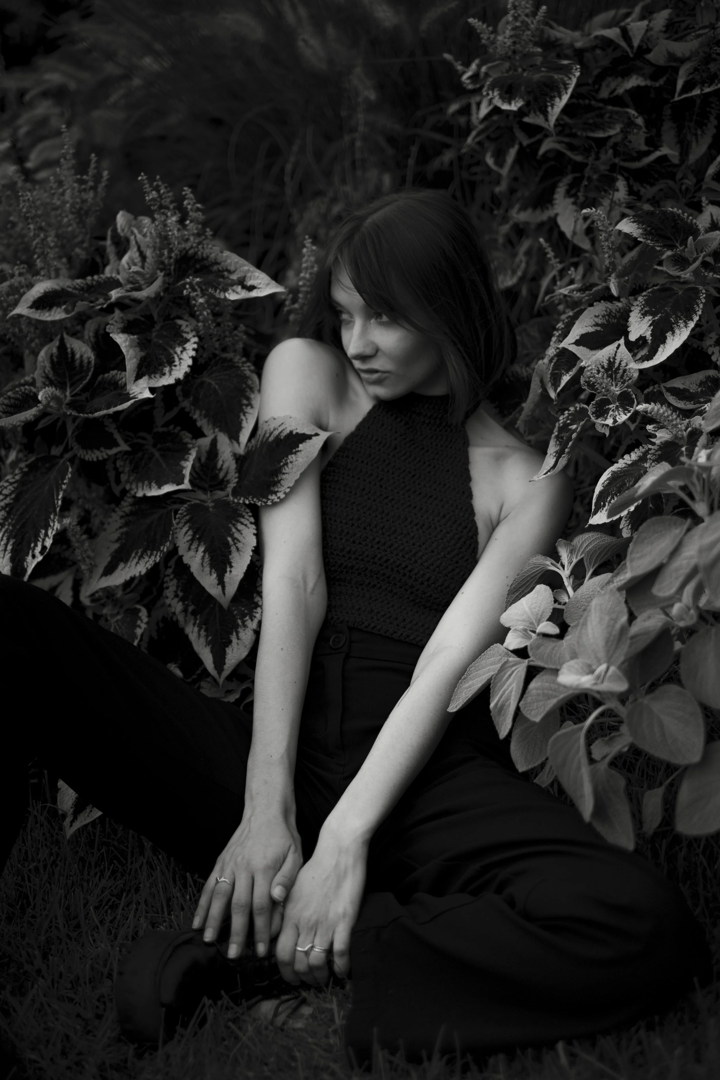 Une jeune femme aux cheveux courts, assise parmi des feuilles et des plantes, regardant sur le côté avec un léger sourire.