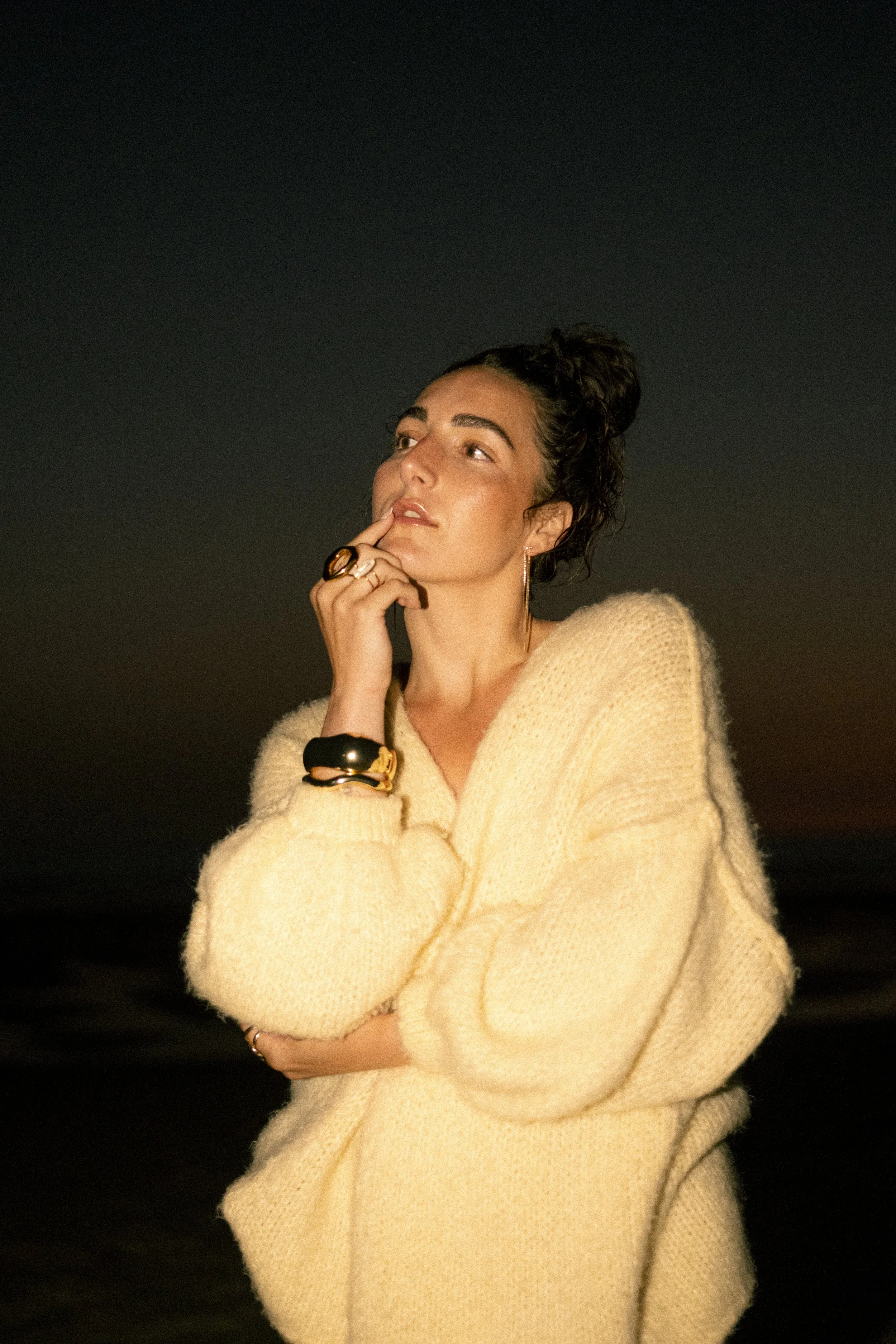 Une femme en pull beige regarde pensivement vers la gauche, avec le doigt sur la bouche, en arrière-plan noir.
