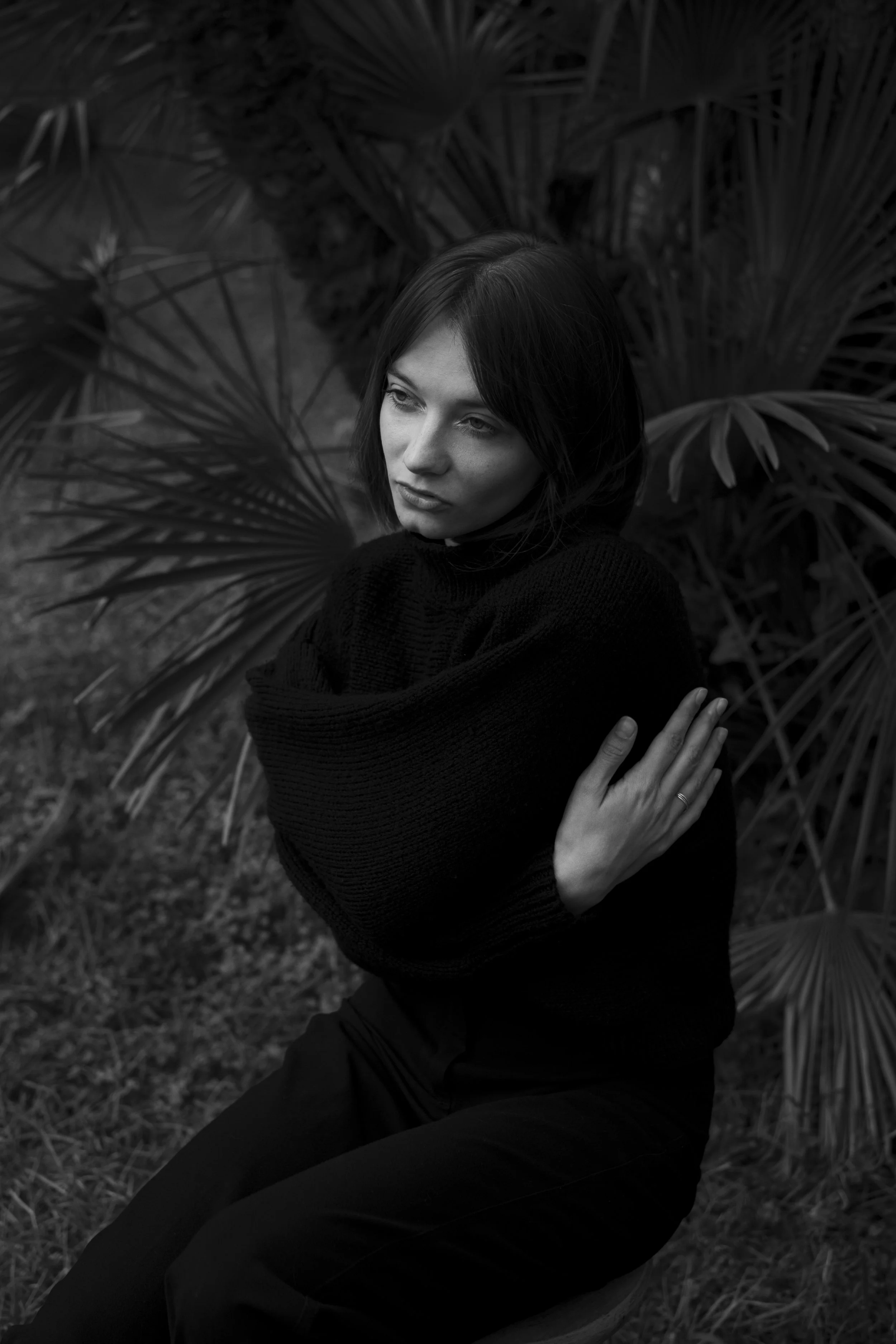 Une femme aux cheveux courts, en pull noir, se tient parmi des feuilles de palmier, regardant légèrement sur le côté dans une photo en noir et blanc.
