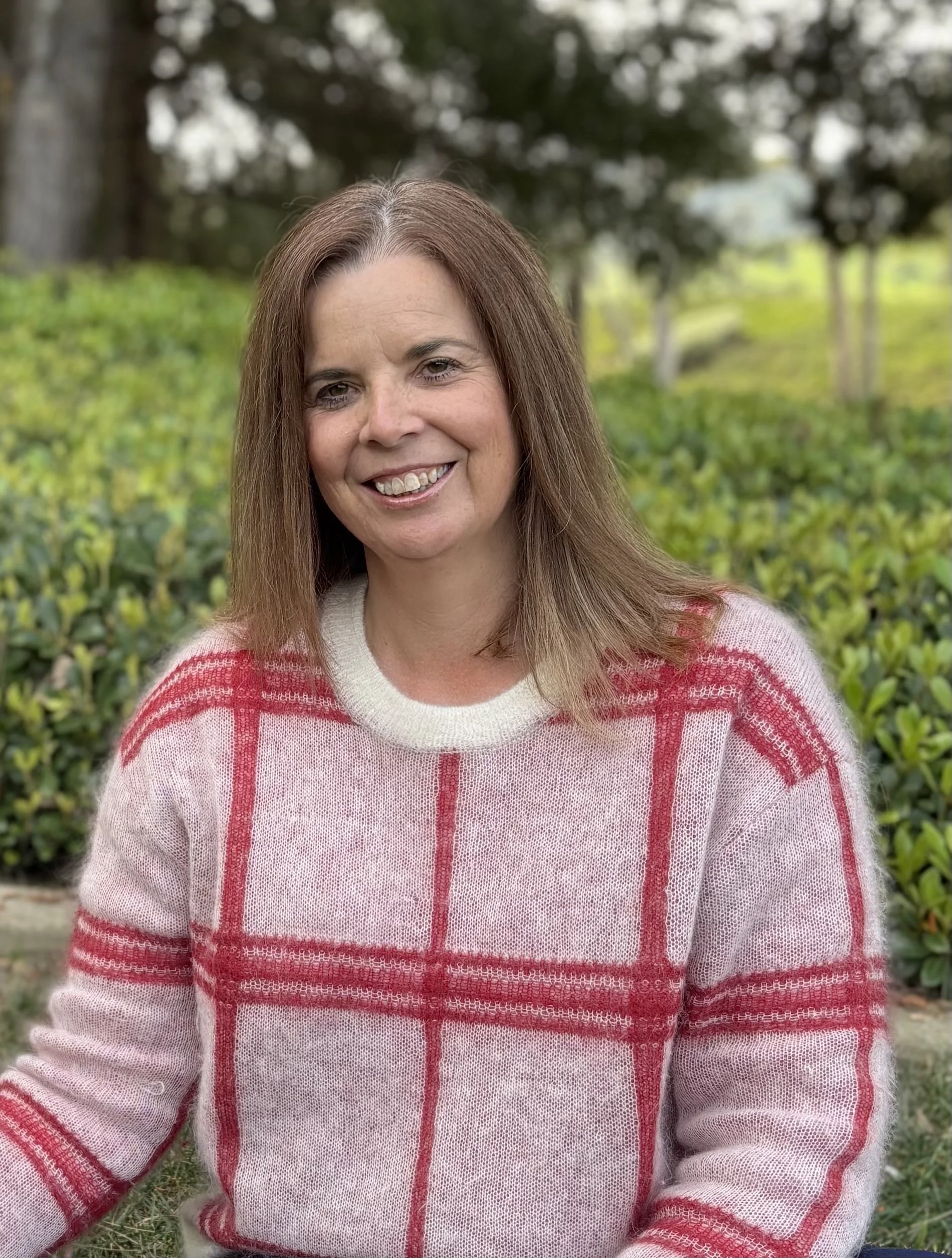 A woman with shoulder-length brown hair, smiling, wearing a cream-colored sweater with red plaid pattern, sitting outdoors with greenery and trees in the background.