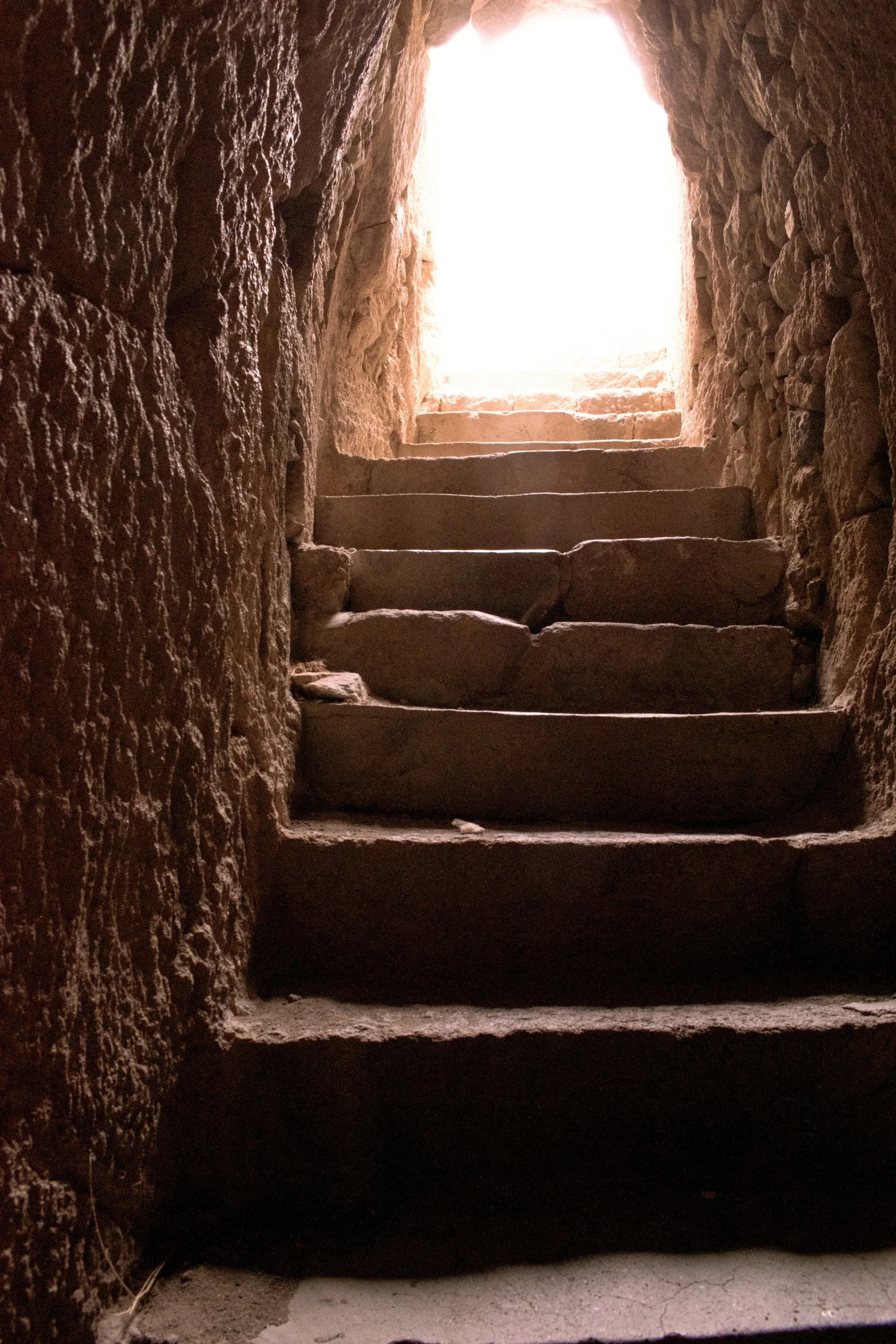Stone stairs leading up to a bright opening at the top, with sunlight shining through.