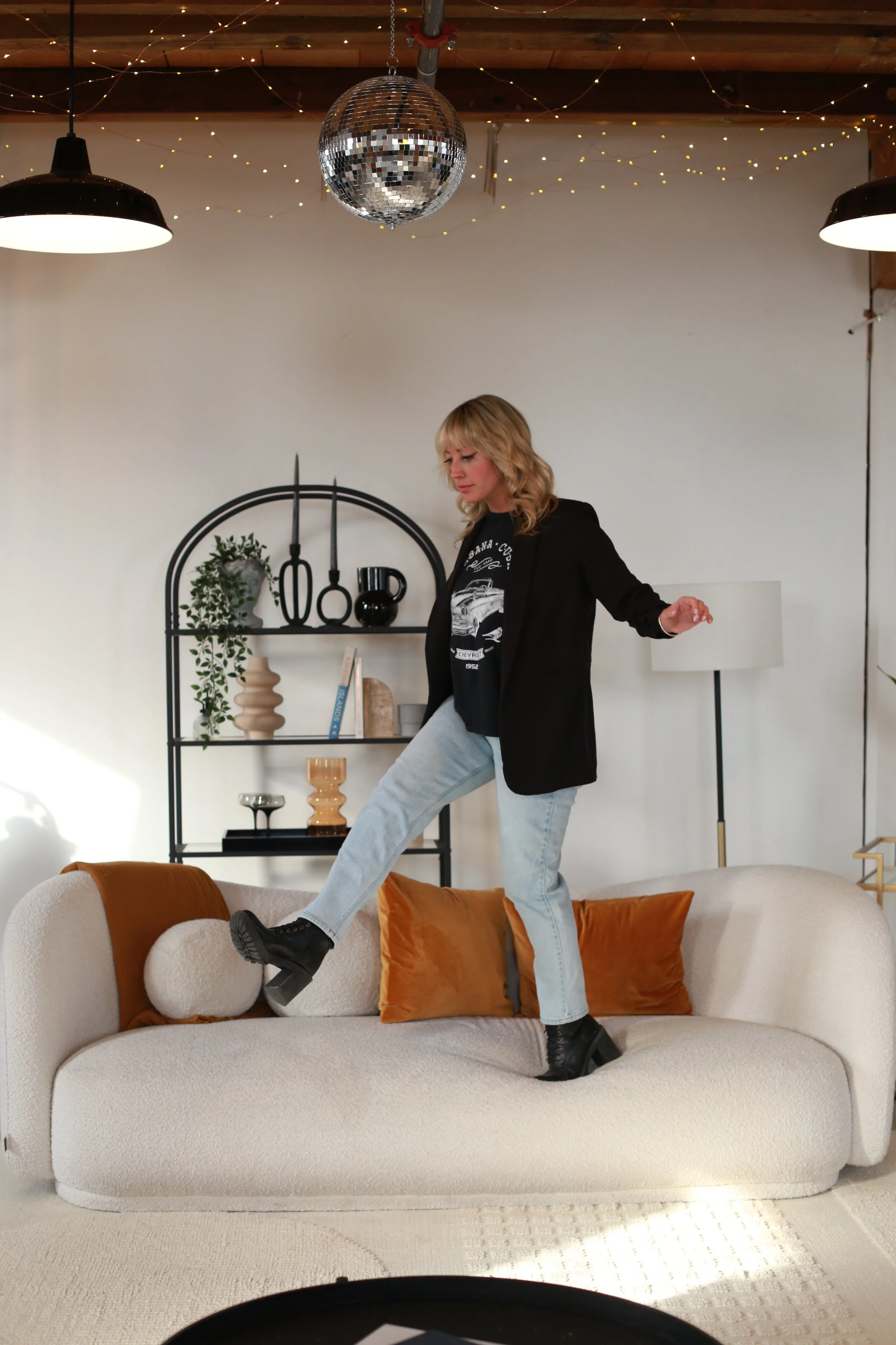 Woman in black blazer, graphic T-shirt, light jeans, and platform boots standing on a white sofa beneath a disco ball and string lights