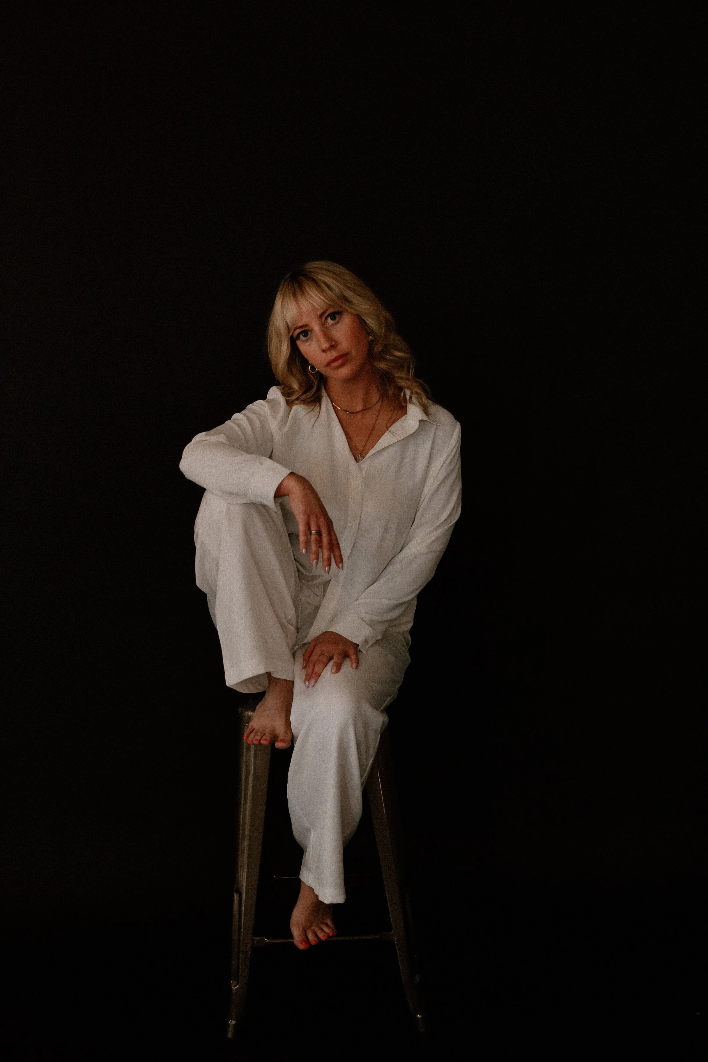 Blonde woman in a white outfit sitting barefoot on a chair against a black background