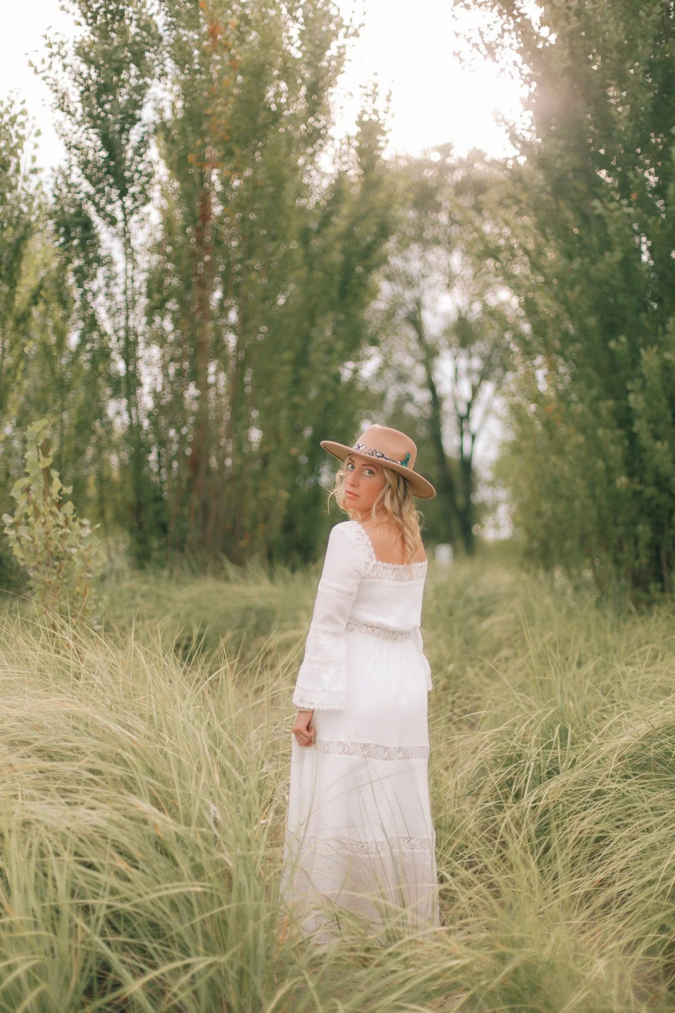 Blonde woman in a white lace dress and wide-brimmed hat standing in tall grass at the edge of a sunlit forest