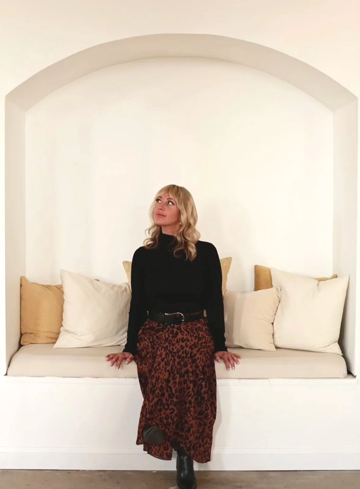 Blonde woman wearing a black top and leopard-print skirt sitting on a white built-in window seat with pillows