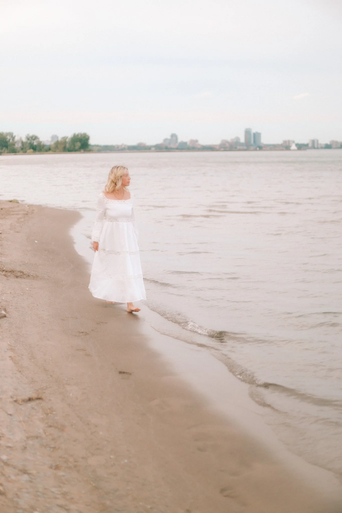 Woman in a white dress walking barefoot along a sandy lakeshore with a distant city skyline
