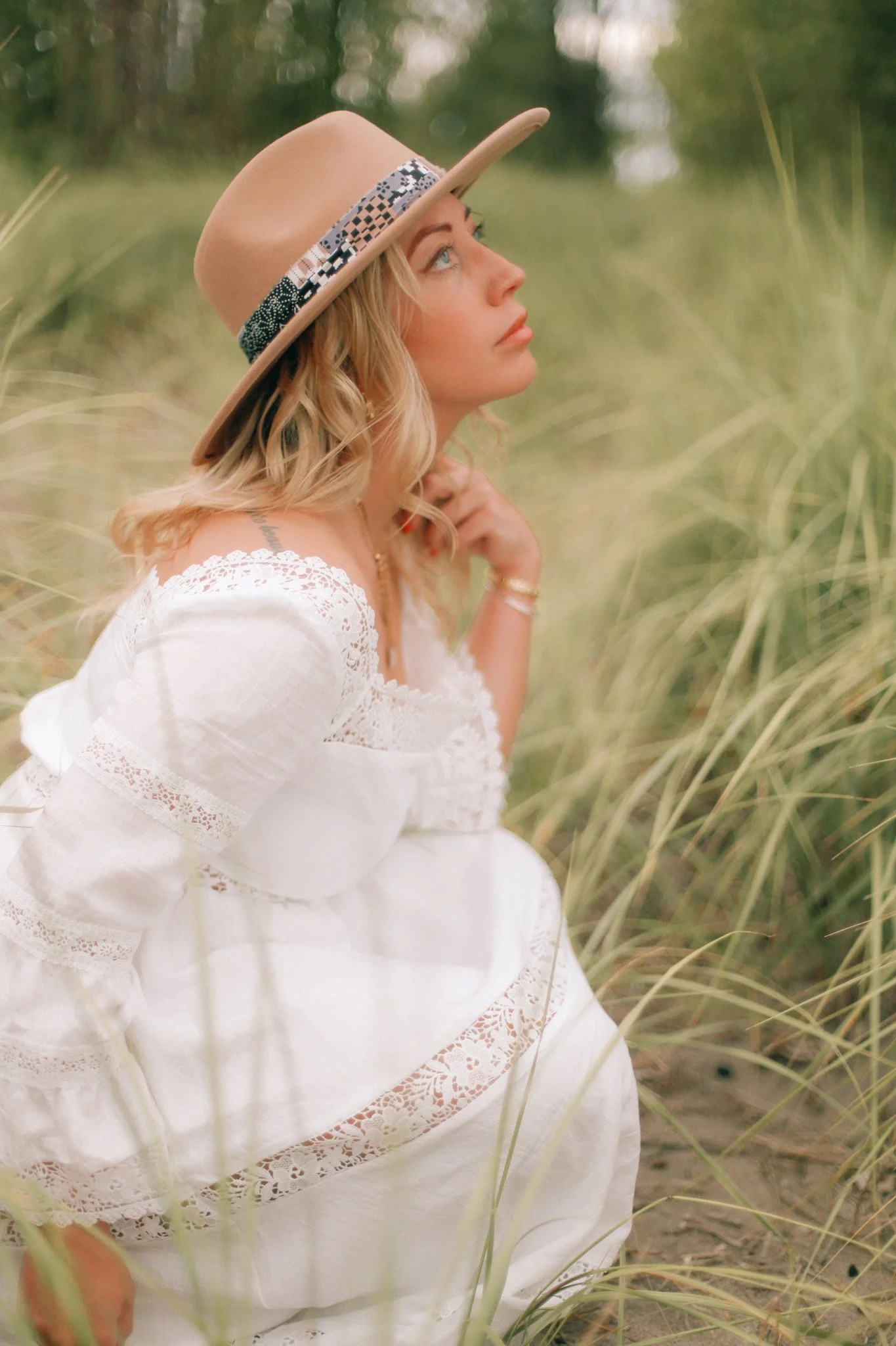 Blonde woman in a patterned straw hat and white lace-trimmed dress sitting among tall grasses
