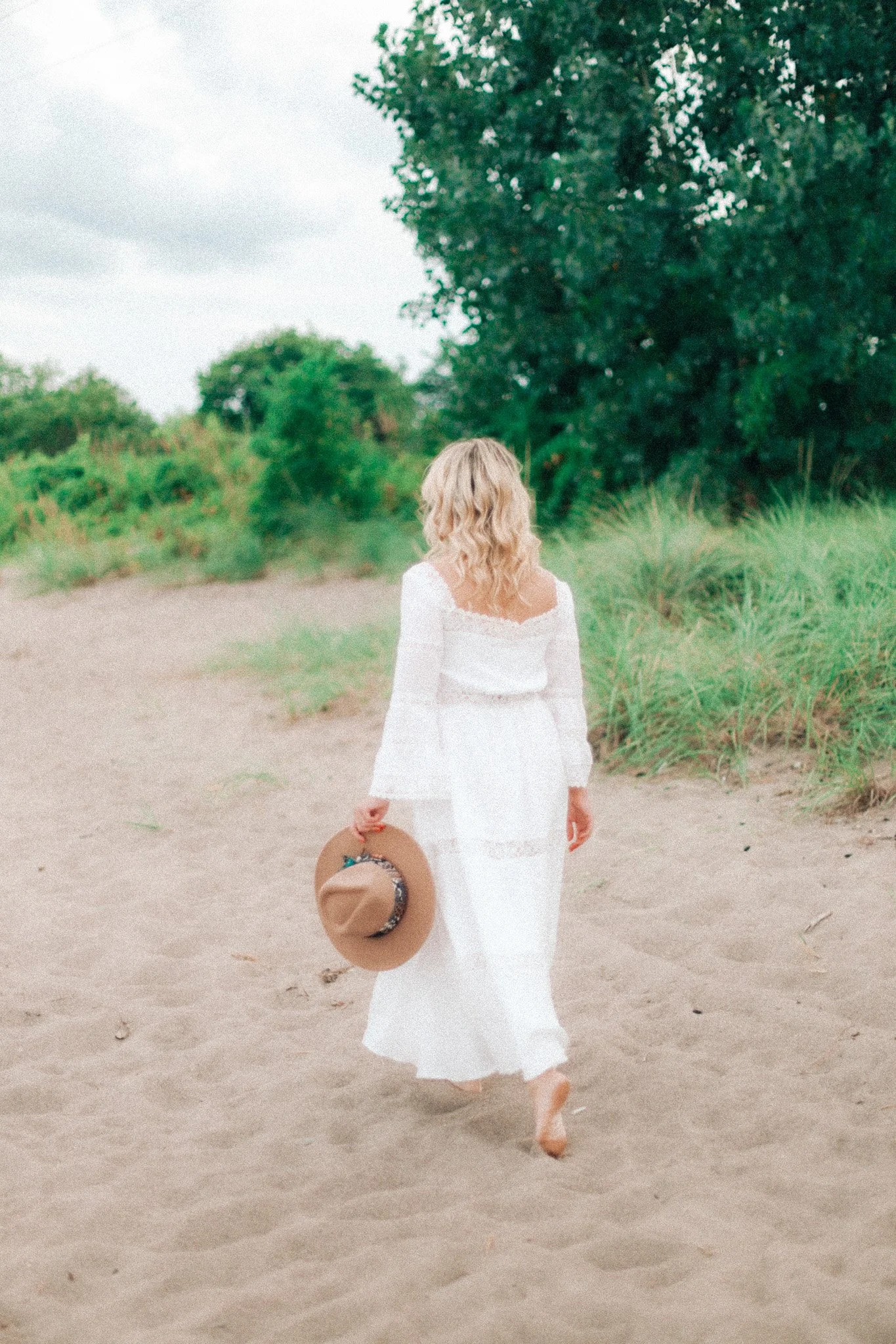 Woman in a white dress walking along a sandy path, holding a wide-brimmed hat, surrounded by green trees and grass