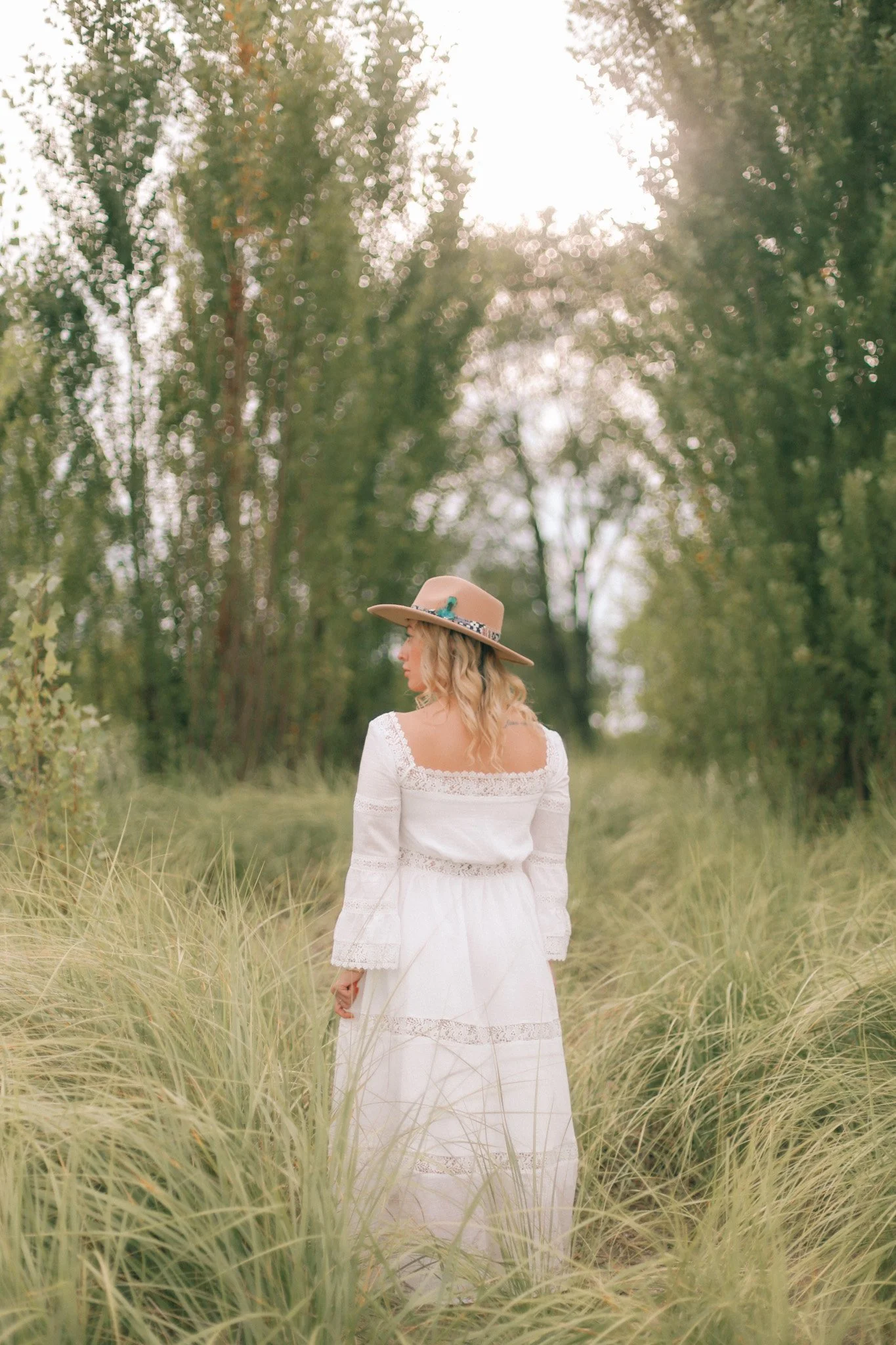 Woman in a white lace dress and wide-brimmed hat standing in tall grass in a sunlit forest