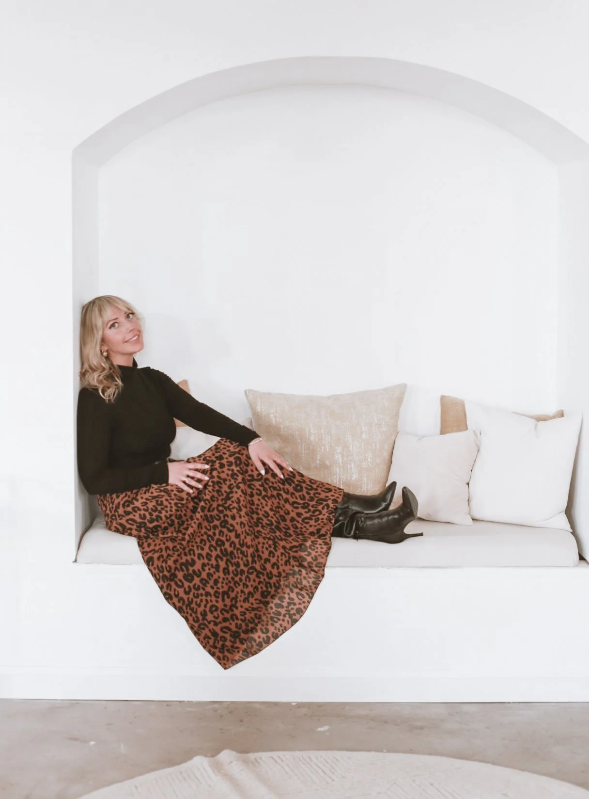 Woman in a black top and leopard-print skirt reclining on a white built-in sofa with pillows and black boots