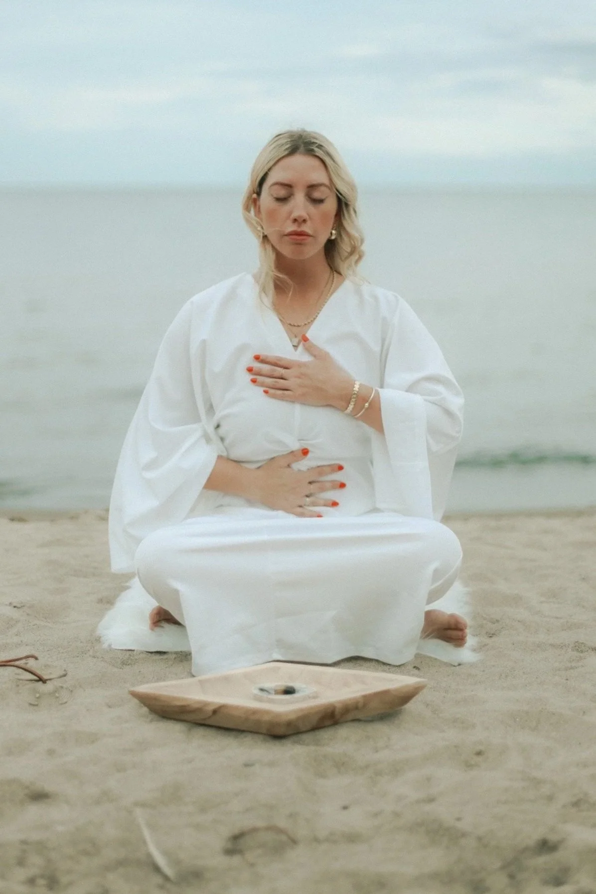 Woman in white clothing meditating cross-legged on a beach with hands on her chest and abdomen