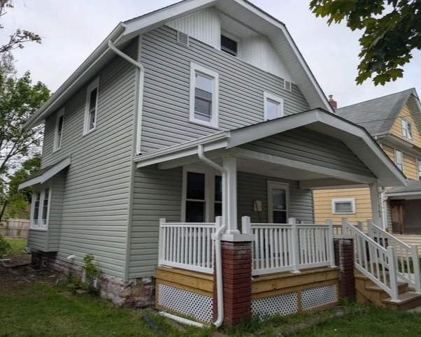 A two-story light gray house with a front porch, white railing, brick and wood supports, located in a suburban neighborhood. After picture