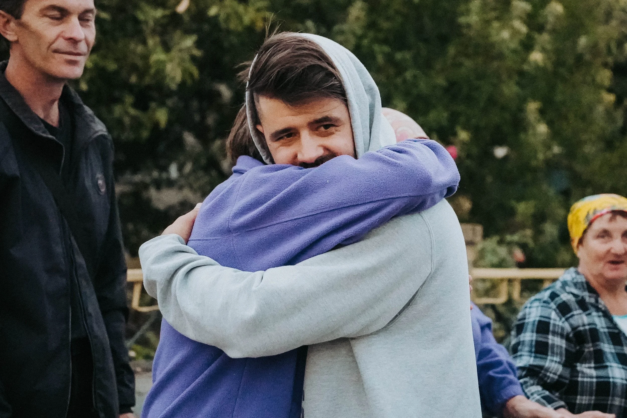 Two young men are hugging outdoors among a group of people, with another person wearing a headscarf visible in the background.
