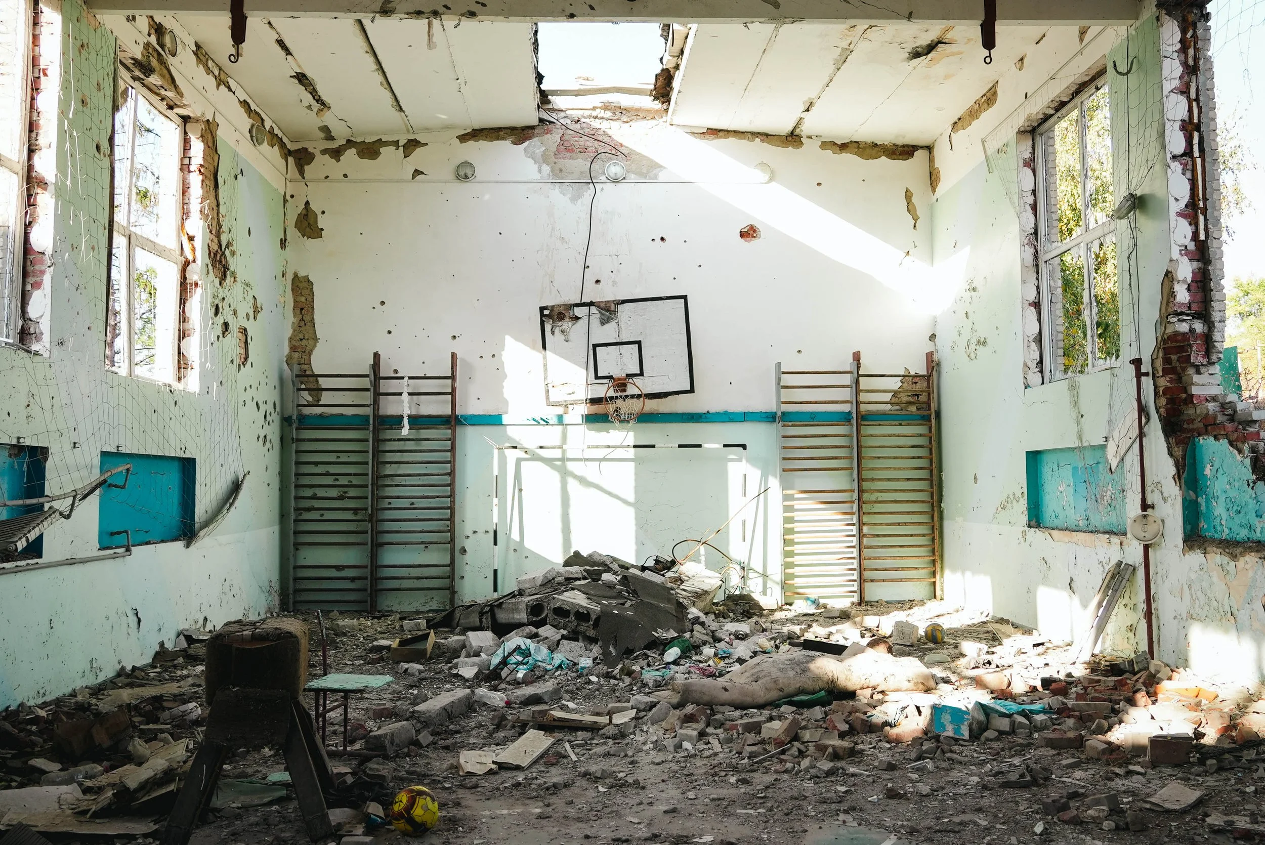 An abandoned, destroyed indoor gymnasium with damaged walls, scattered debris, and a basketball hoop on the wall.