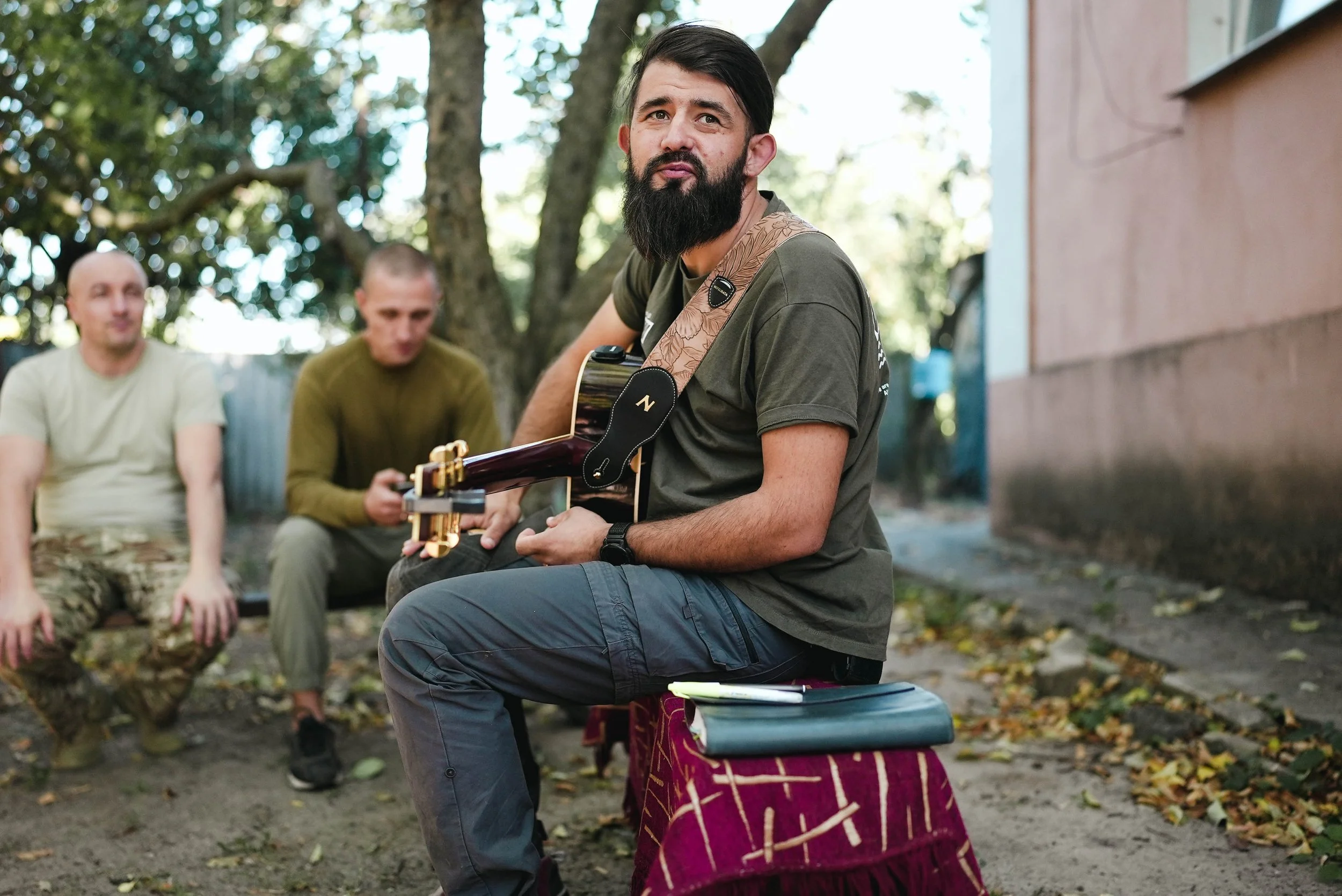 A man with a beard playing guitar while sitting outdoors on a red cloth-covered stool, with three other men sitting behind him in a garden with trees and a building in the background.
