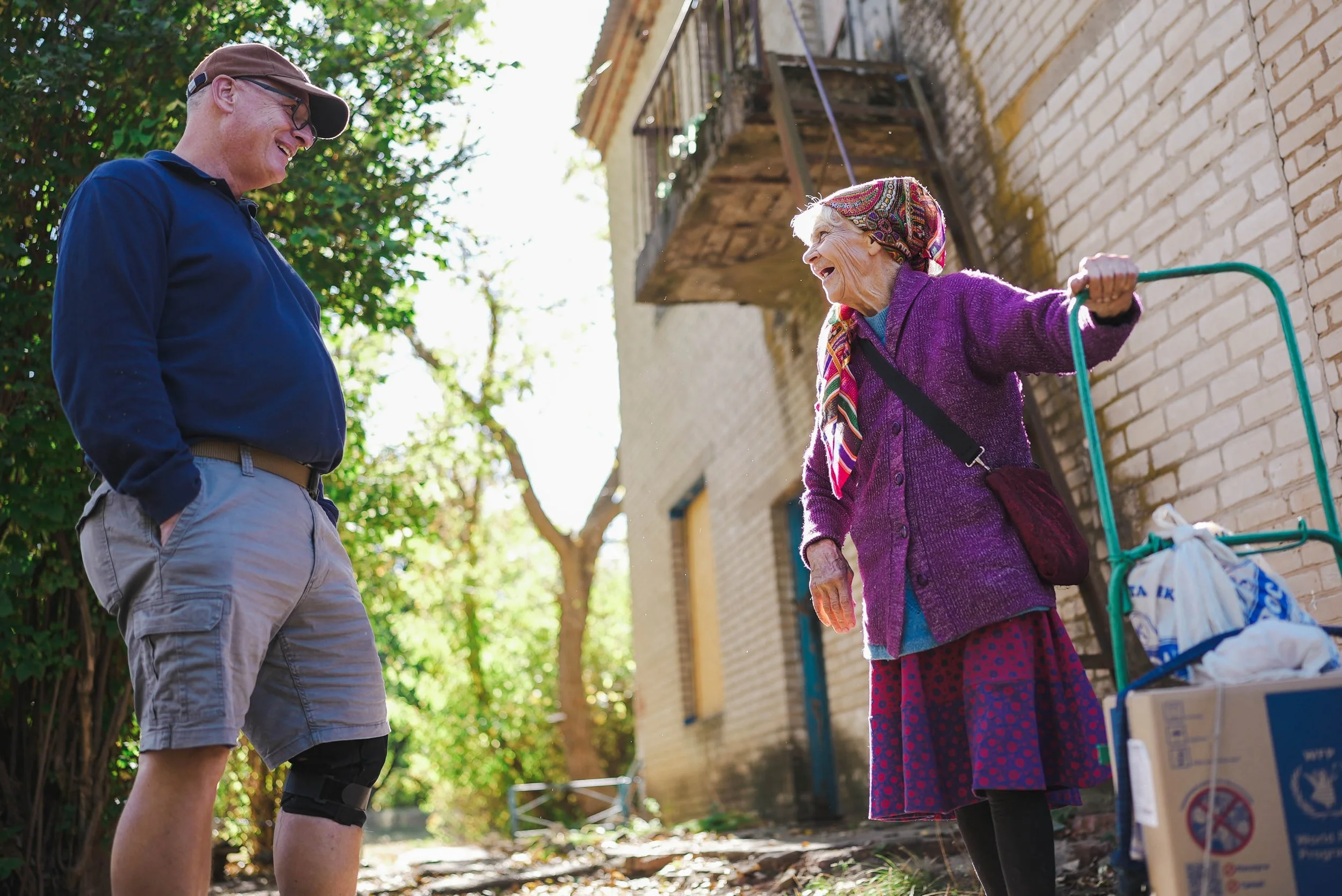 An elderly woman and a middle-aged man smiling and talking outdoors on a sunny day. The woman is wearing a purple cardigan, a colorful headscarf, and a patterned skirt, with a walker nearby. The man is wearing a blue polo shirt, khaki shorts, glasses, and a cap, with a knee brace on his leg.