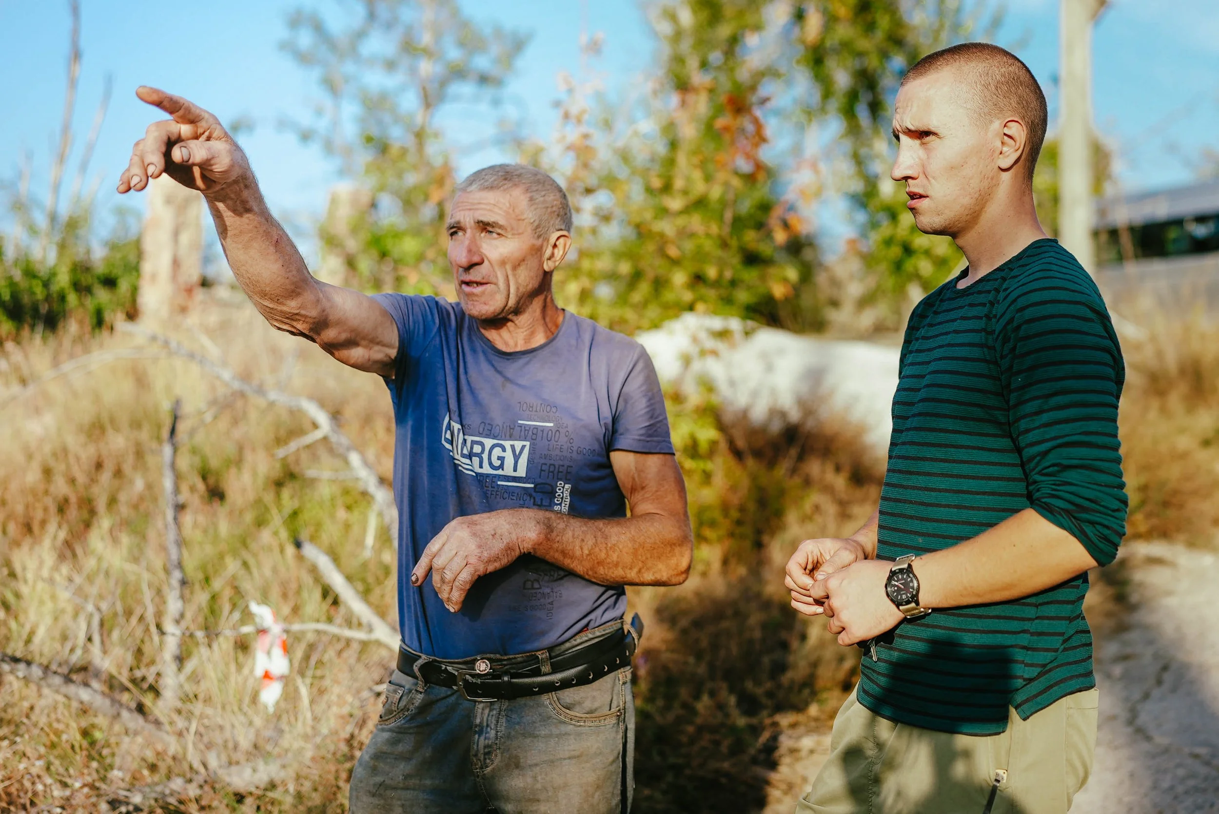 An older man with short gray hair and a younger man with short hair outdoors, with trees and a dirt path in the background. The older man is pointing at something while speaking, and the younger man is listening attentively.