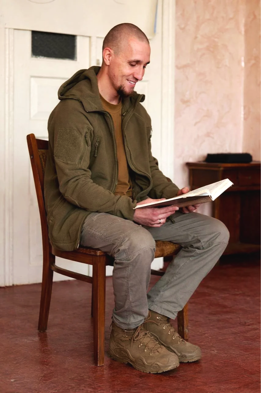 A man with a shaved head and beard, dressed in a green tactical jacket, khaki pants, and hiking boots, is sitting on a wooden chair reading a book in a room with a wooden floor, a white door, and a peach-colored wall.