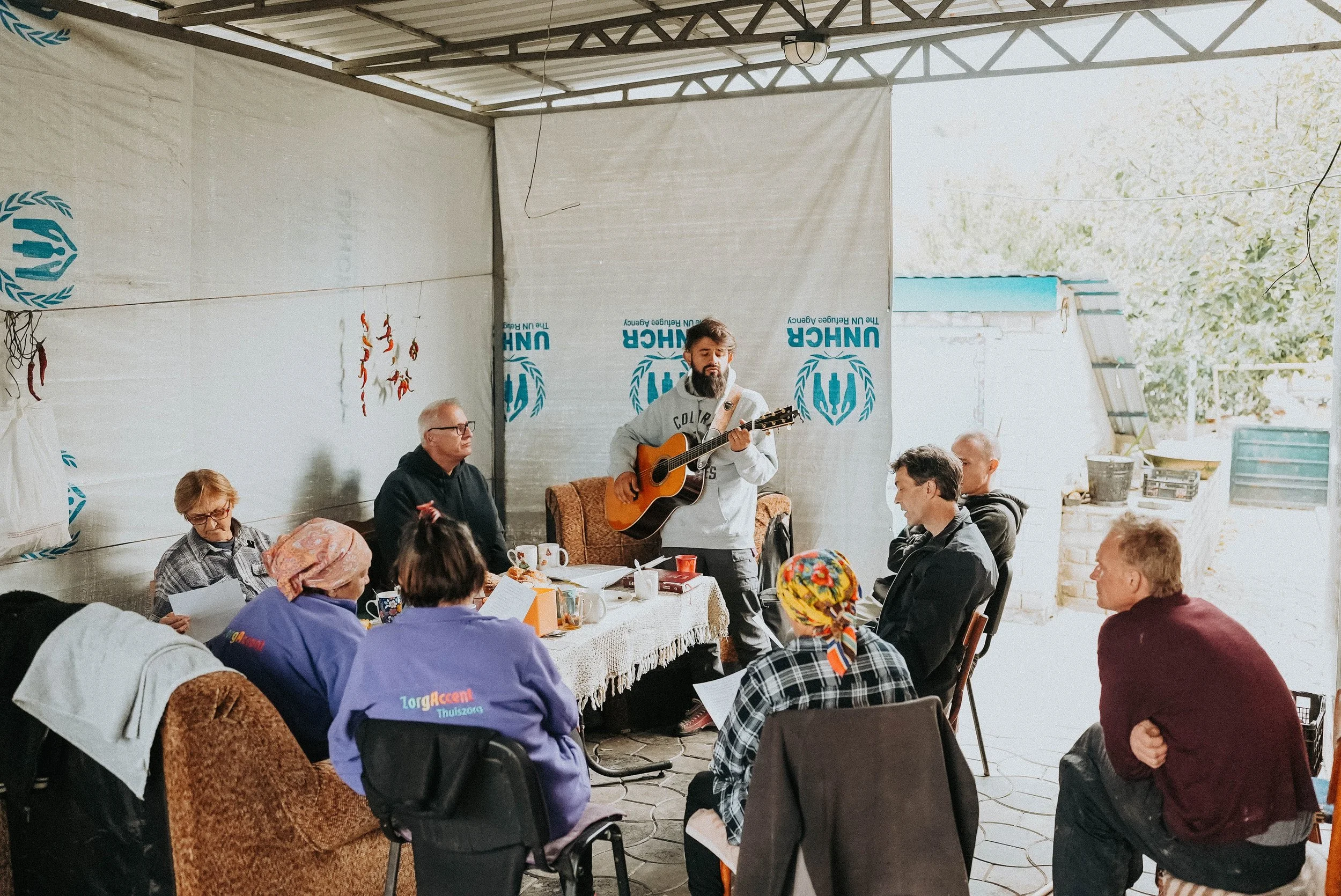 A group of people sitting around a table in a semi-outdoor space, with one man standing and playing guitar, and others singing or listening.