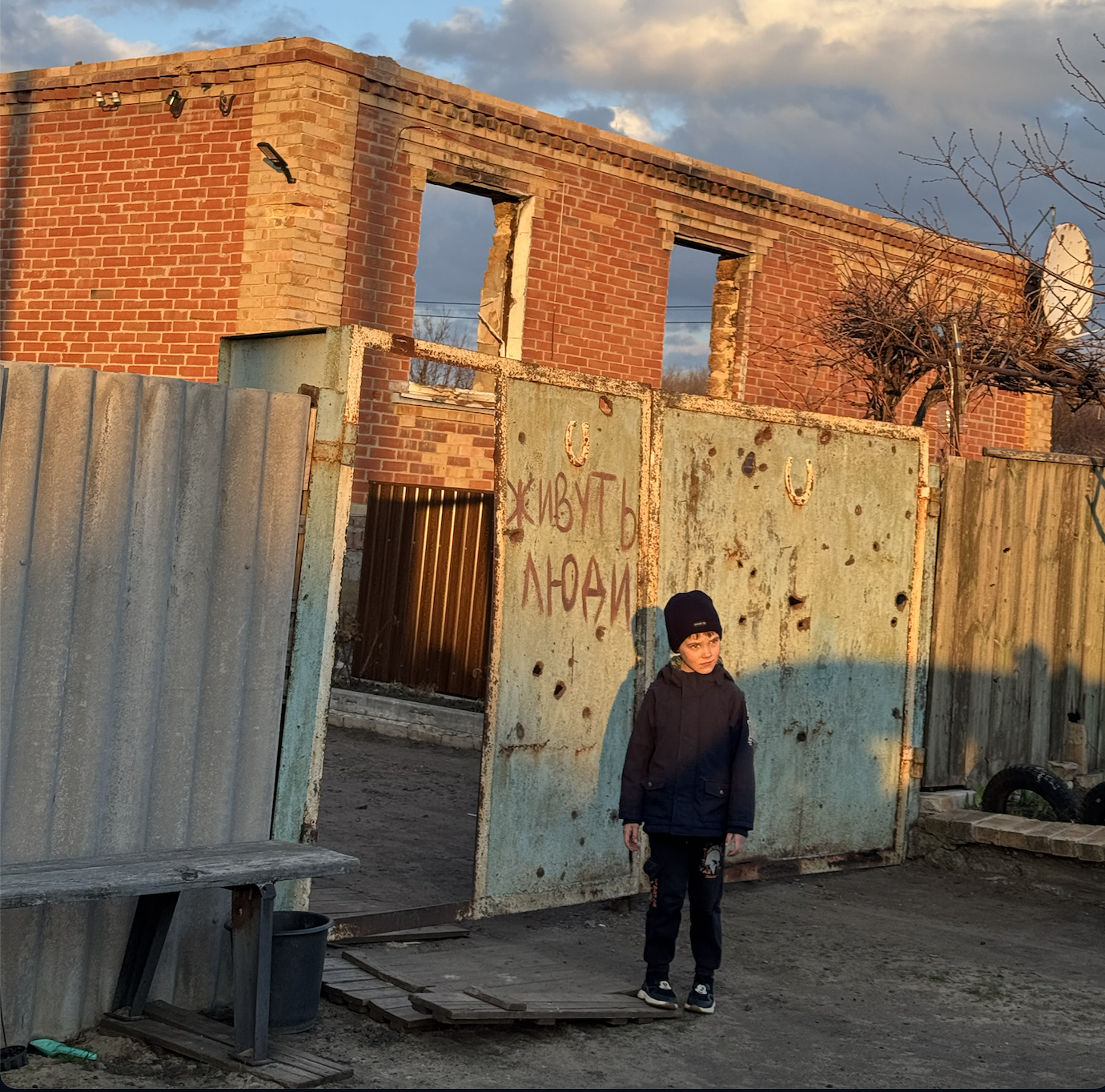 A young boy in dark clothes and a black beanie standing in front of a rusted metal gate with a partially exposed brick building behind it. The building has no windows and appears abandoned. The gate has graffiti in Russian, and the scene is bathed in warm sunlight during what seems to be late afternoon or early evening.
