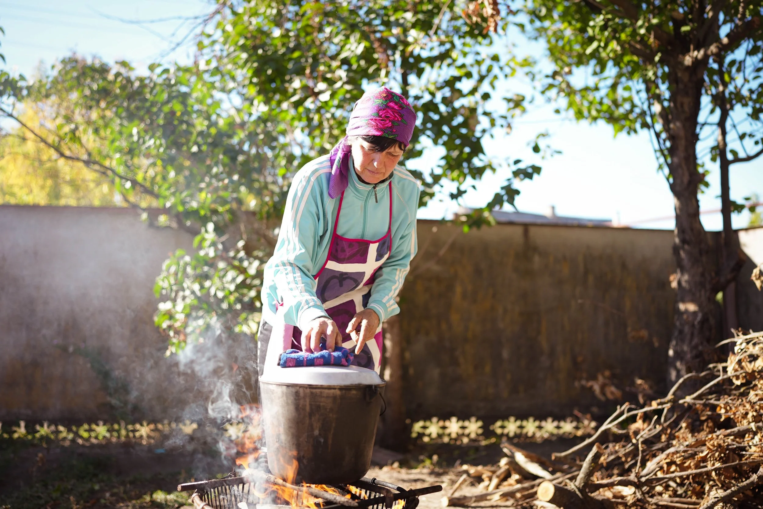 An elderly woman wearing a headscarf and apron, standing outdoors by a tree, cooking or tending to a pot over an open fire.