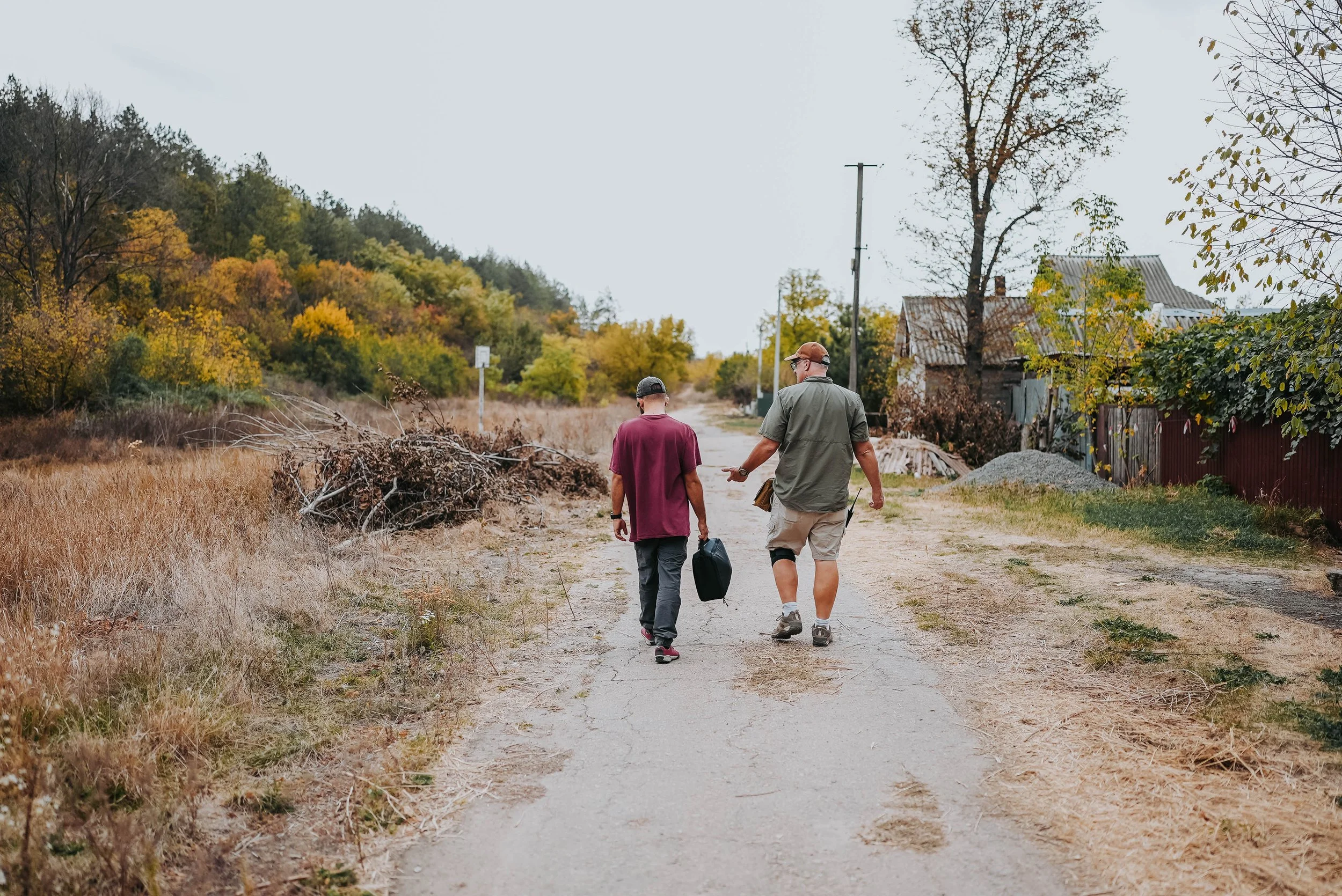 Two men walking down a rural dirt path in autumn, one carrying a bag, the other gesturing, with trees and old houses on either side.