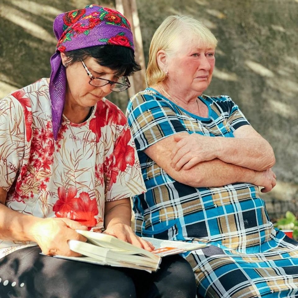 Two older women sitting outdoors; one wearing glasses, a purple headscarf with red roses, and a floral shirt, looking through a book or magazine; the other with arms crossed, wearing a blue and brown plaid shirt, looking away with a serious expression.