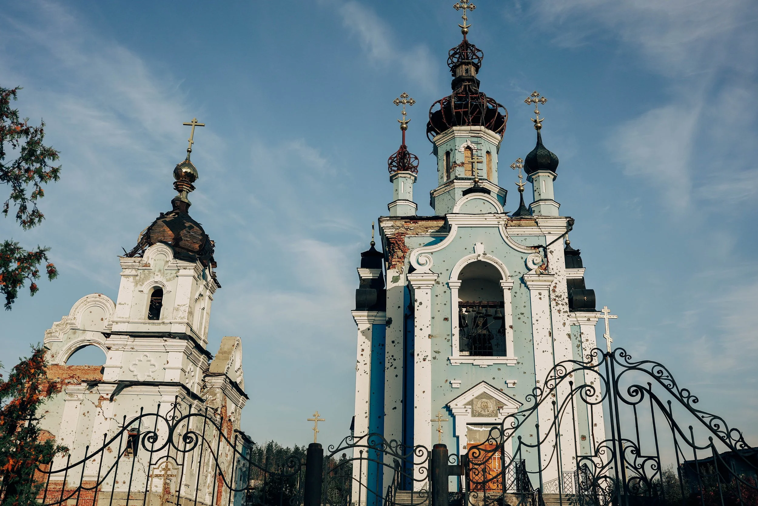 An old, damaged Orthodox church with peeling light blue and white paint, multiple onion domes topped with crosses, surrounded by a black wrought iron fence, under a partly cloudy sky.