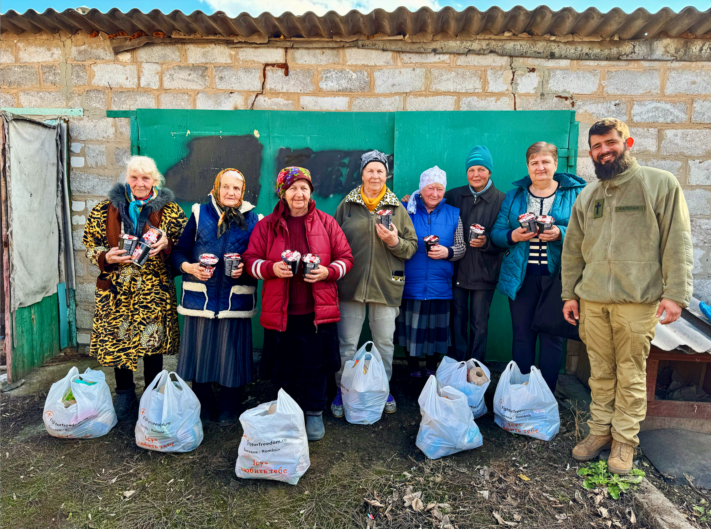 A group of nine people, mostly elderly women and one man, standing outdoors in front of a brick and green door wall, holding containers of food. They are dressed warmly, with some women wearing headscarves and colorful jackets. Plastic bags filled with supplies are on the ground in front of them. The setting appears to be a rural or modest residential area.