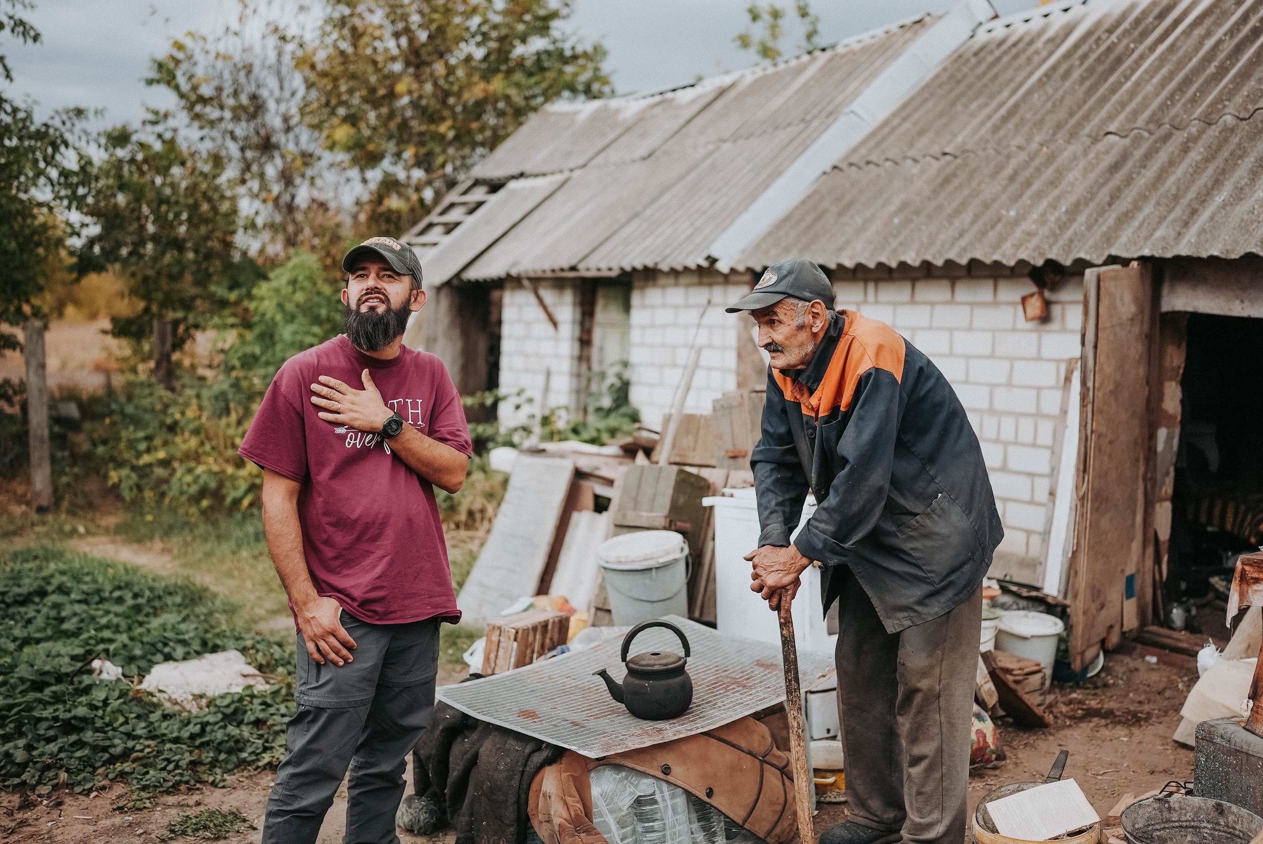 Two men stand outside a rustic building with a corrugated metal roof; one man is an elderly with a cane, the other is a younger man with his hand over his chest.