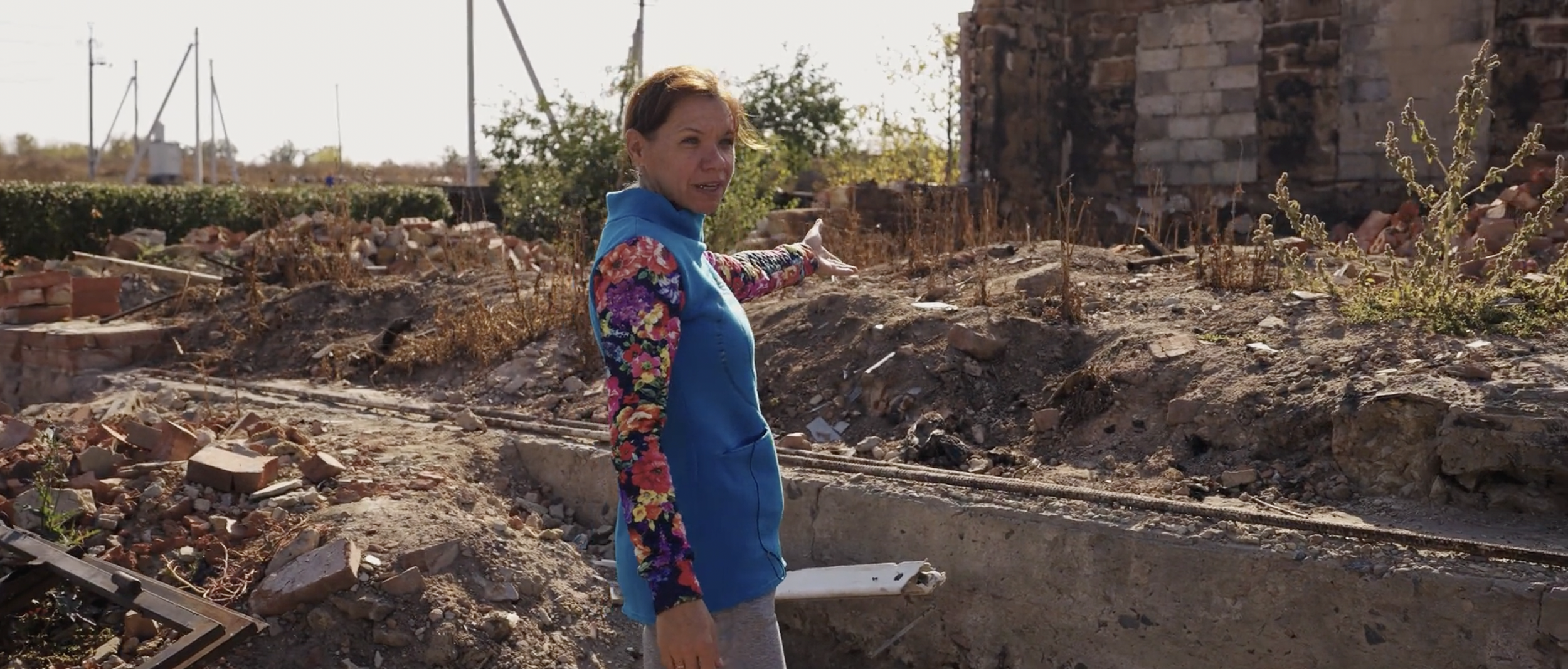 A woman in a bright blue jacket with floral sleeves points at a damaged, collapsed area with rubble and debris, indicating destruction or construction, outdoors during daytime.