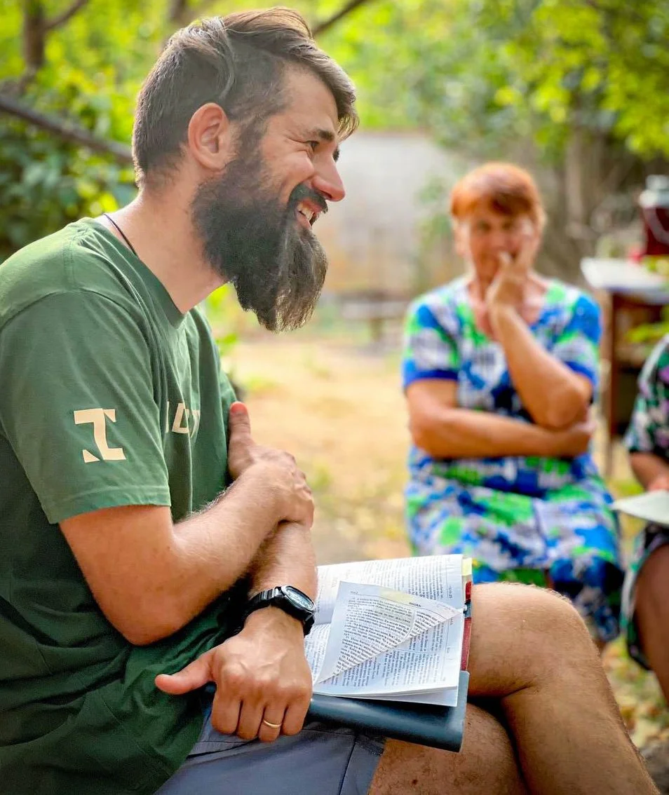 A man with a beard, wearing a green t-shirt and a watch, reading a book outdoors in a lush green garden while laughing. In the background, an older woman with short red hair is sitting, smiling, and resting her chin on her hand.