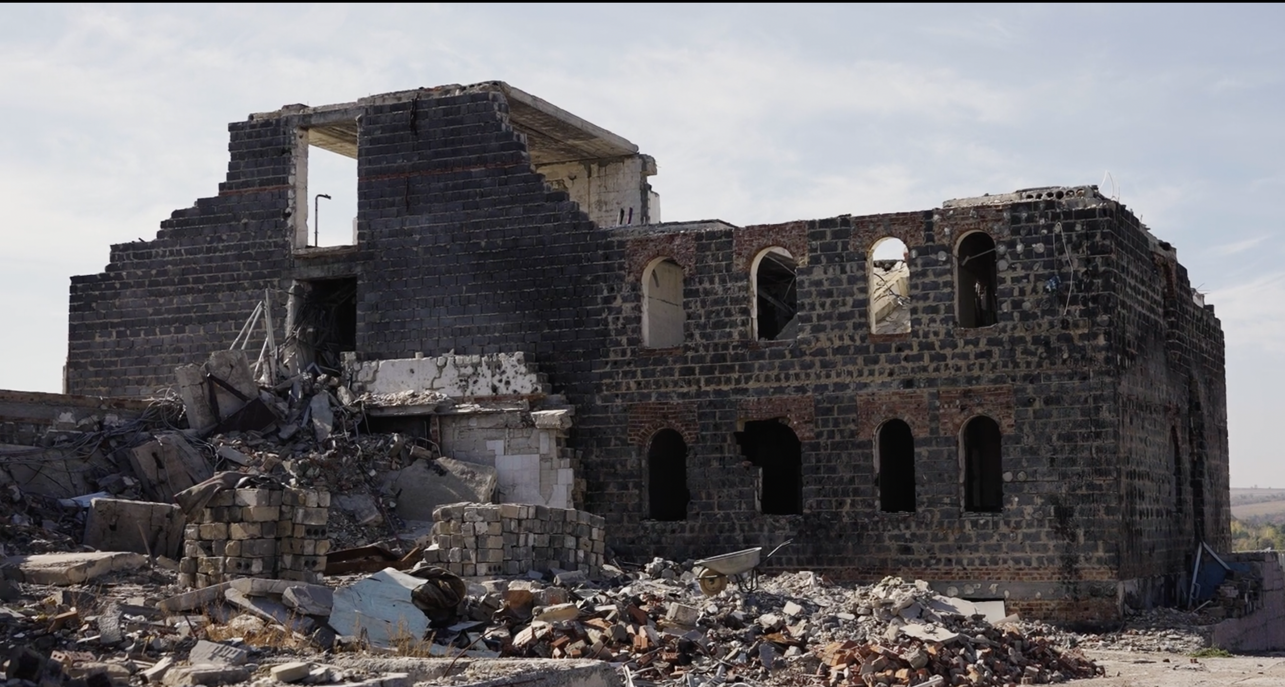 Ruined black brick building with broken windows and debris in front, under a partly cloudy sky.