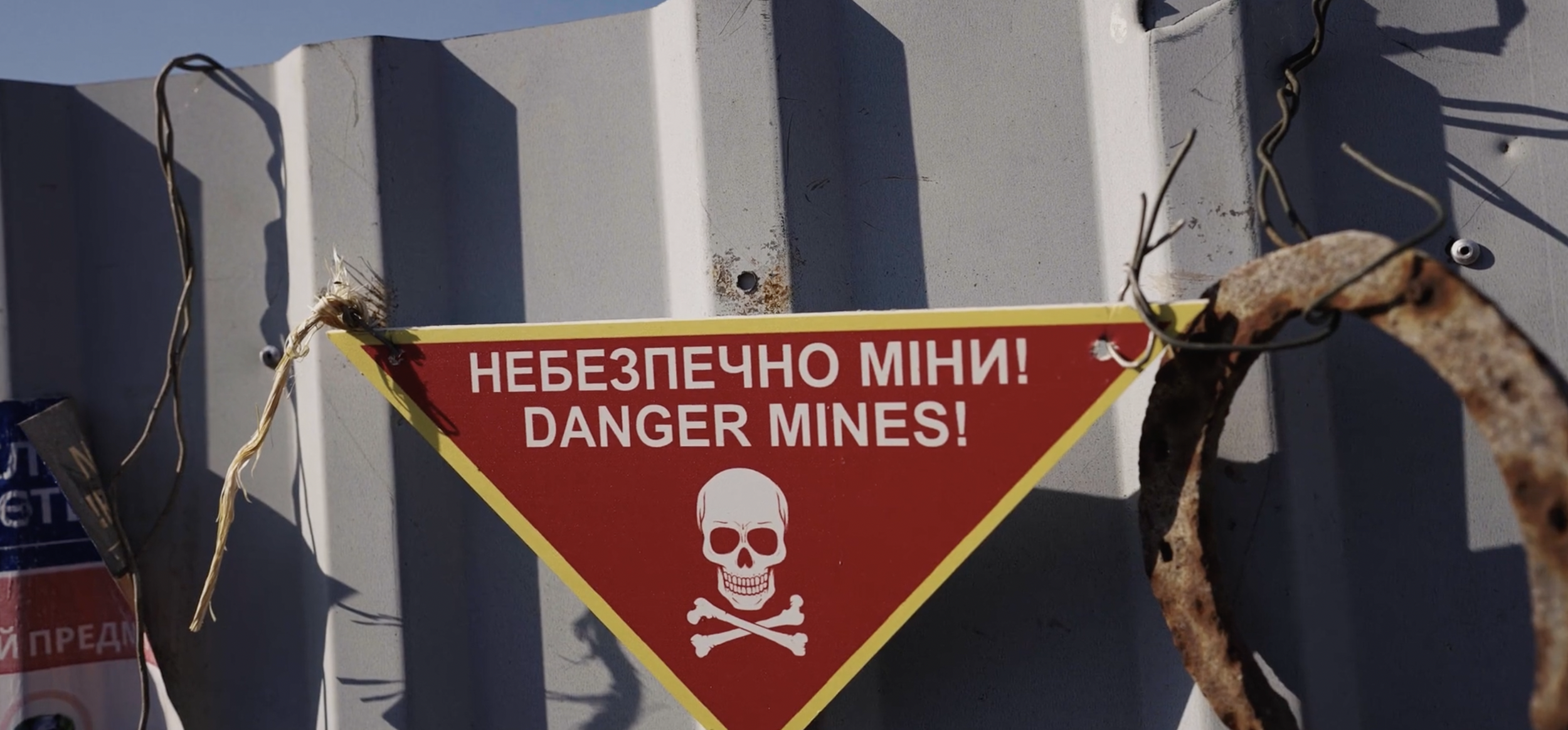 A warning sign with a red background and yellow border that reads "Danger Mines!" in English and Cyrillic script, with a skull and crossbones symbol below the text, attached to a gray metal surface with rust and wires.
