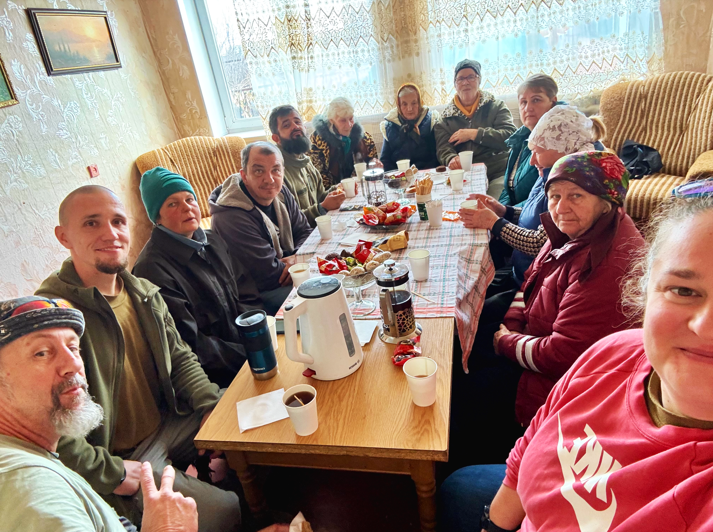 A group of 13 people sitting around a dining table inside a cozy room, enjoying a meal and drinks. There are snacks, a electric kettle, cups, and a tablecloth with a plaid pattern. The room has floral wallpaper, a window with lace curtains, and framed pictures on the wall.