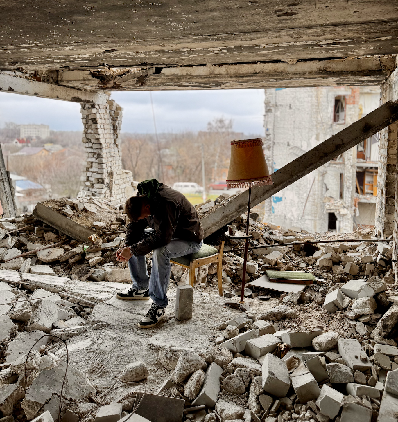 A person sitting on a chair amid rubble and debris in a partially destroyed building, with a table lamp and books nearby, overlooking an urban landscape.