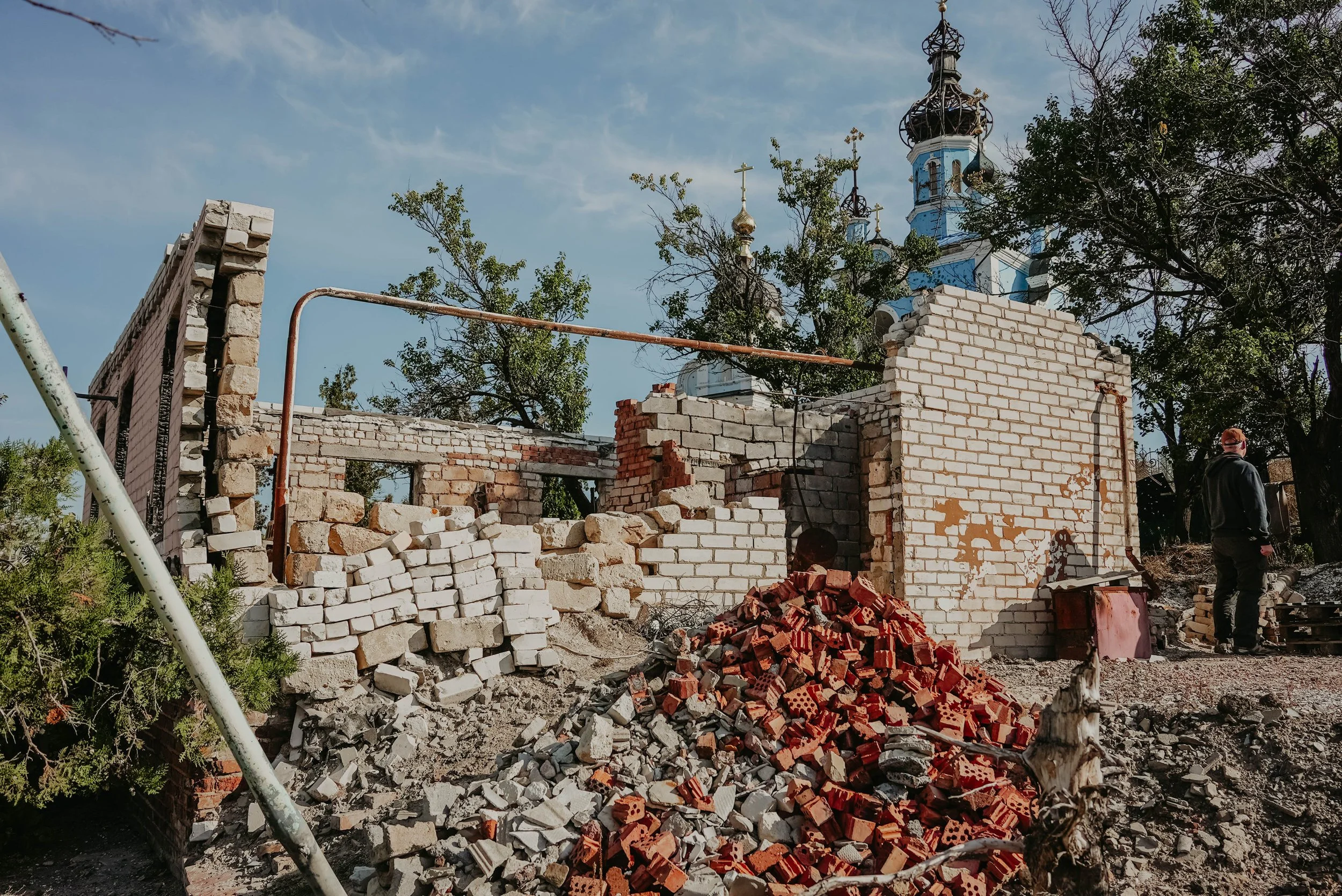 A partially demolished brick building with rubble piled in front, a person standing nearby, and a church with ornate domes and crosses in the background.