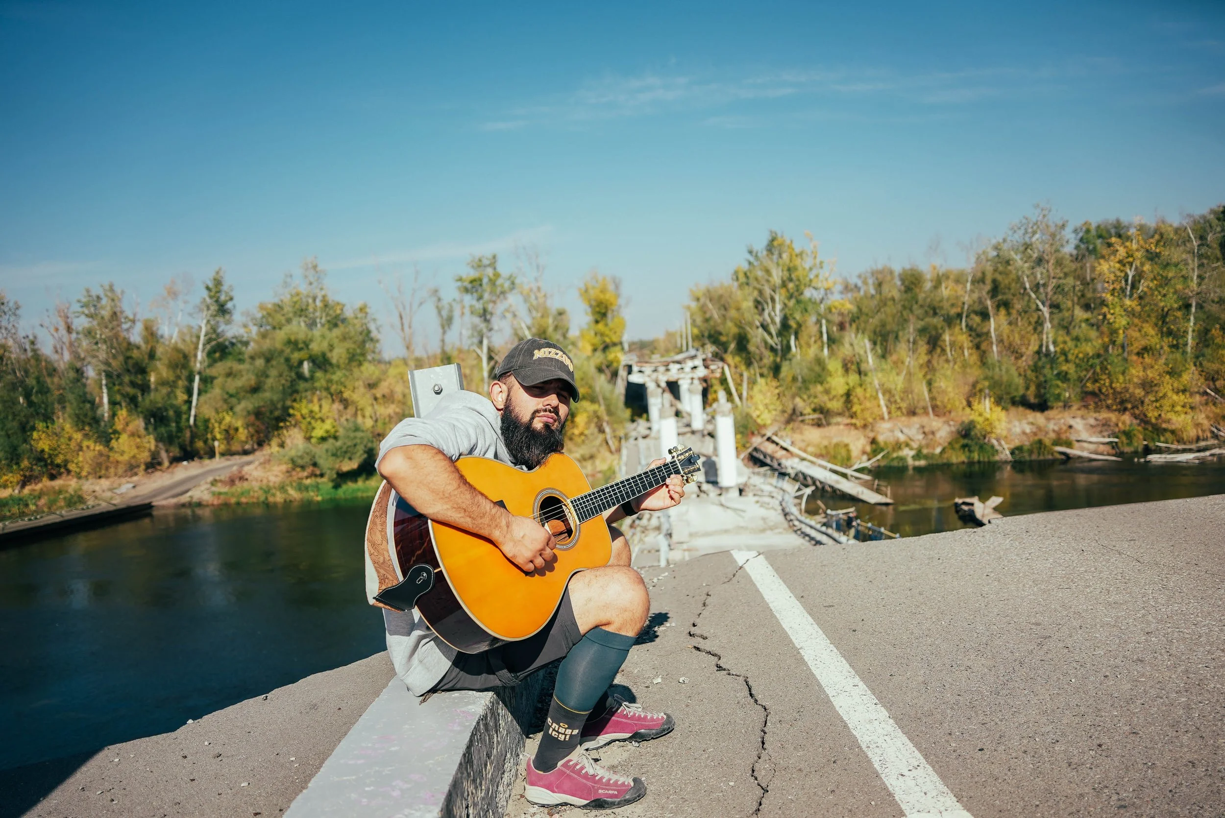 A man with a beard wearing a cap, hoodie, and shorts, sitting on a concrete barrier playing an acoustic guitar near a river with a damaged bridge in the background under a clear blue sky.