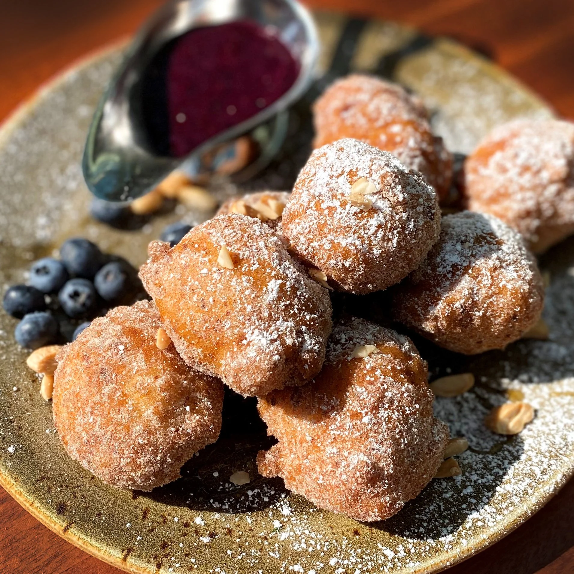 Plate of golden brown deep-fried dough balls dusted with powdered sugar and topped with almond slices, accompanied by fresh blueberries and a glass of berry juice.