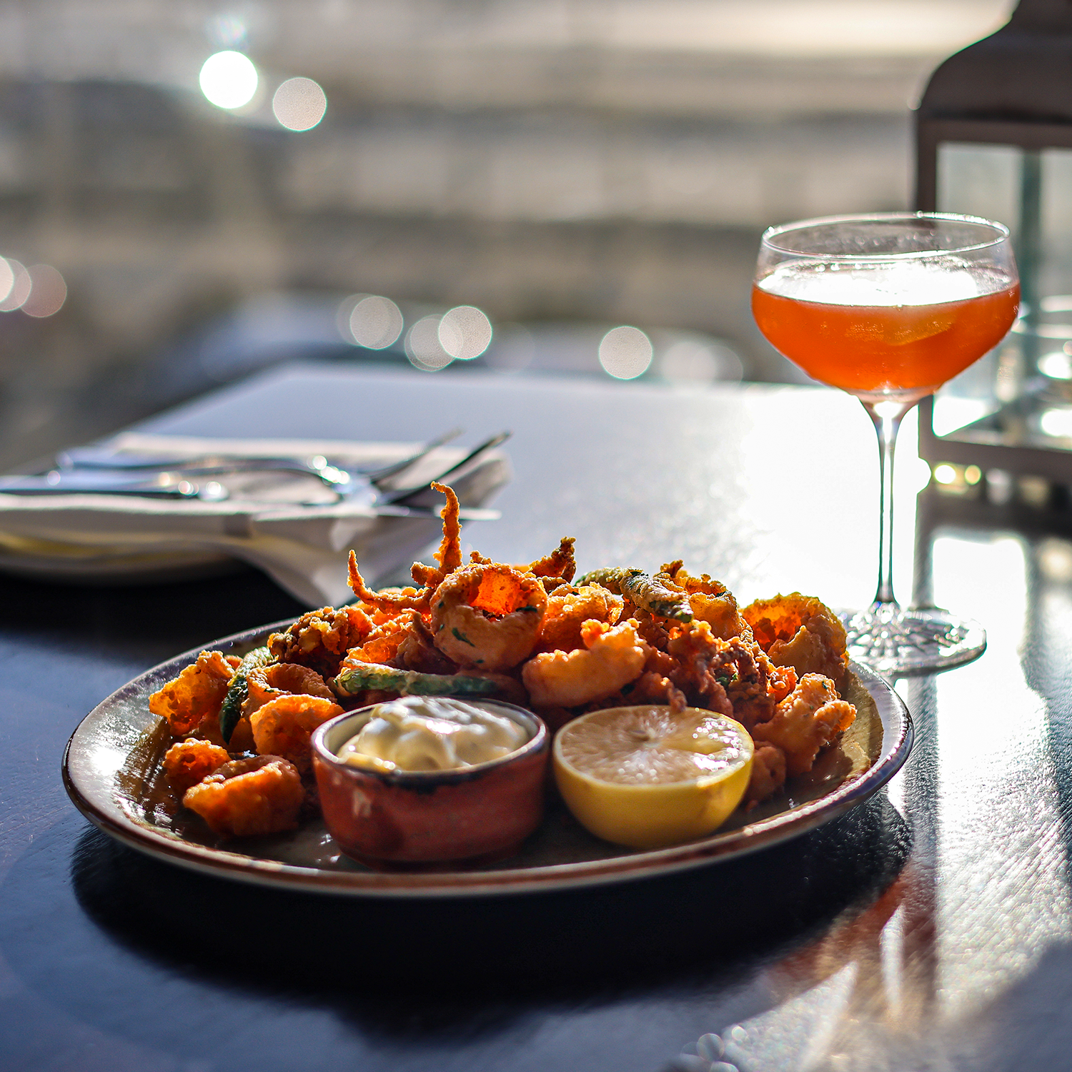 A plate of fried appetizers with lemon and dipping sauces, a glass of red cocktail, and a set of silverware on a table.